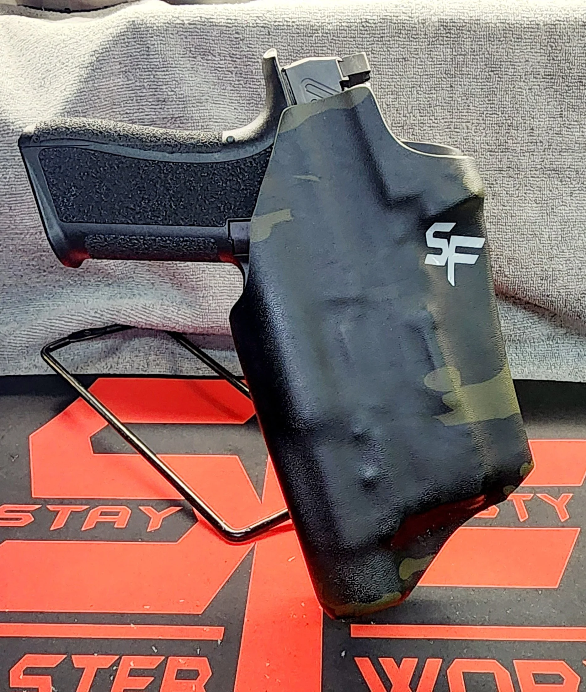 A black firearm with a camouflage cover, placed on a red and black box with the word 'STAY' on it, with a magazine inserted and a mag release button visible.