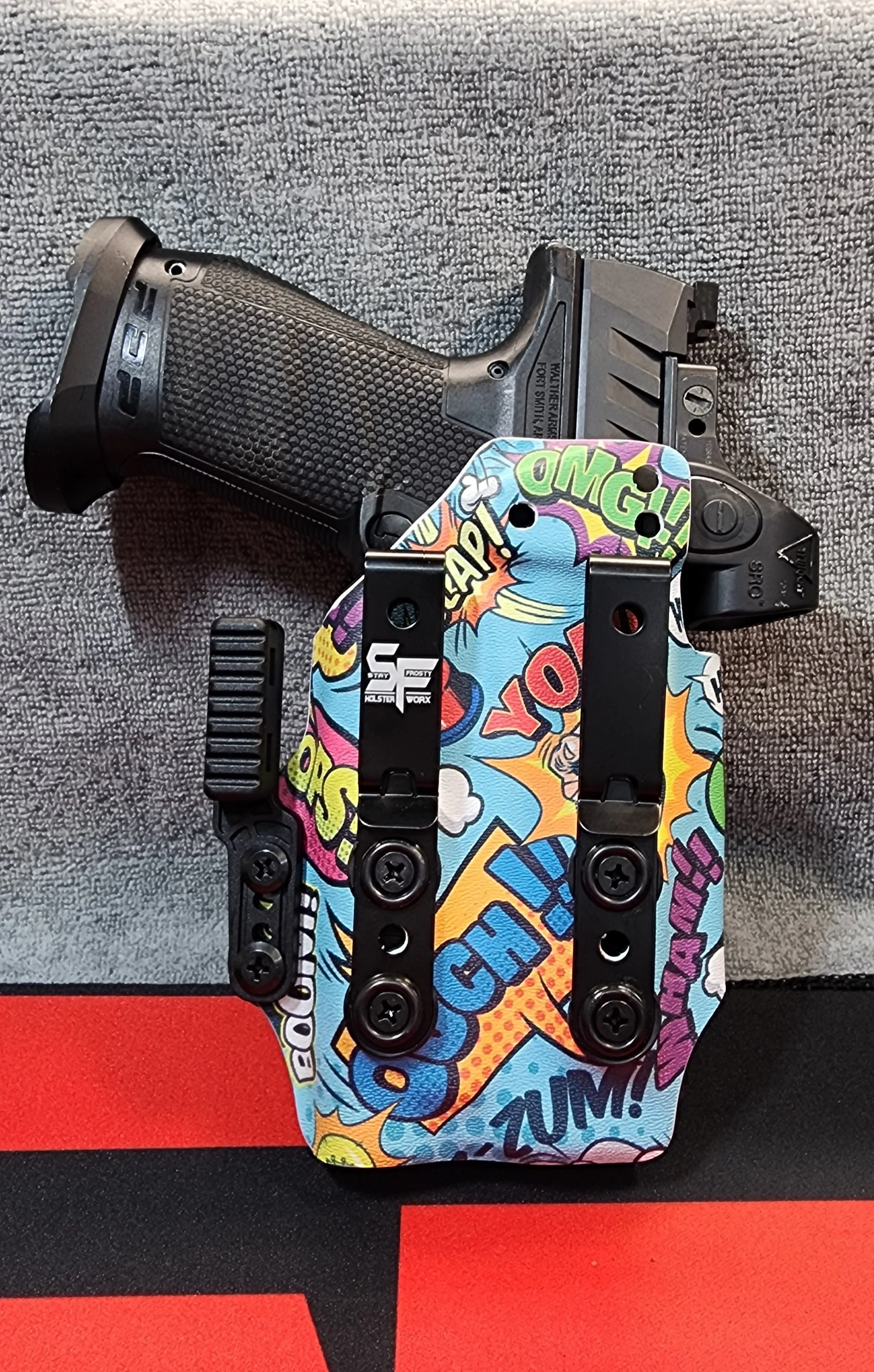 A handgun with a colorful comic book style holster that has words like 'OMG!', 'CRASH!', 'BOOM!', and 'ZUM!' on it, displayed on a gray textured backdrop.