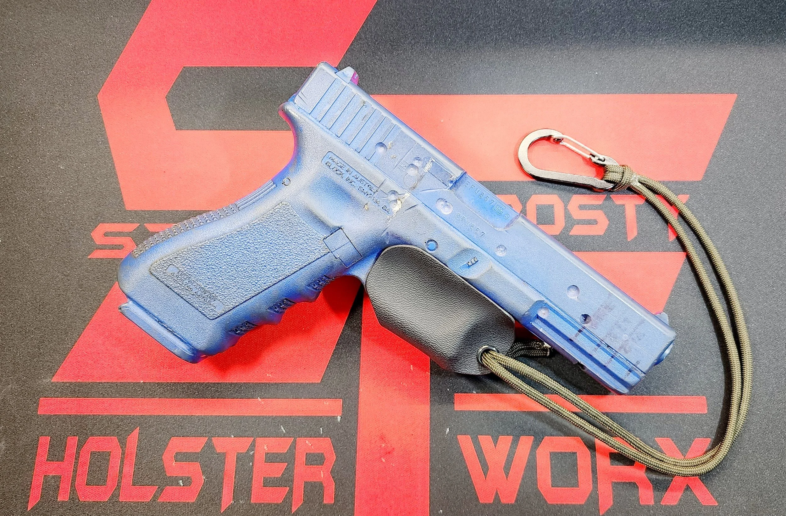 Blue toy gun with black handle on a red and black mat. The mat has the words "Holster Work" and a large stylized "H" and "W" on it. There is a small metal clip attached to the gun with a cord.