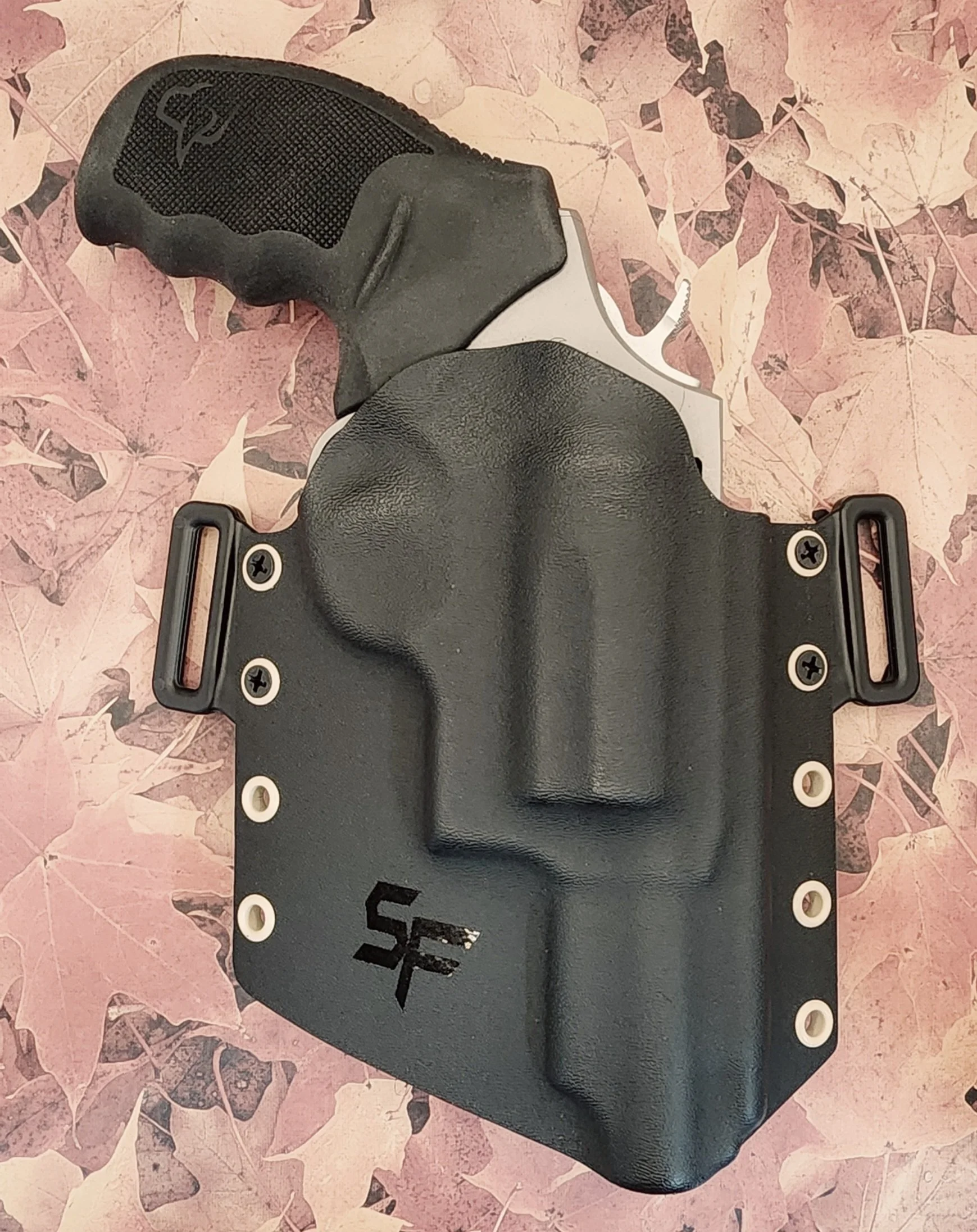 A black firearm holster with a textured grip and a handgun inside, placed on a pink leafy background.