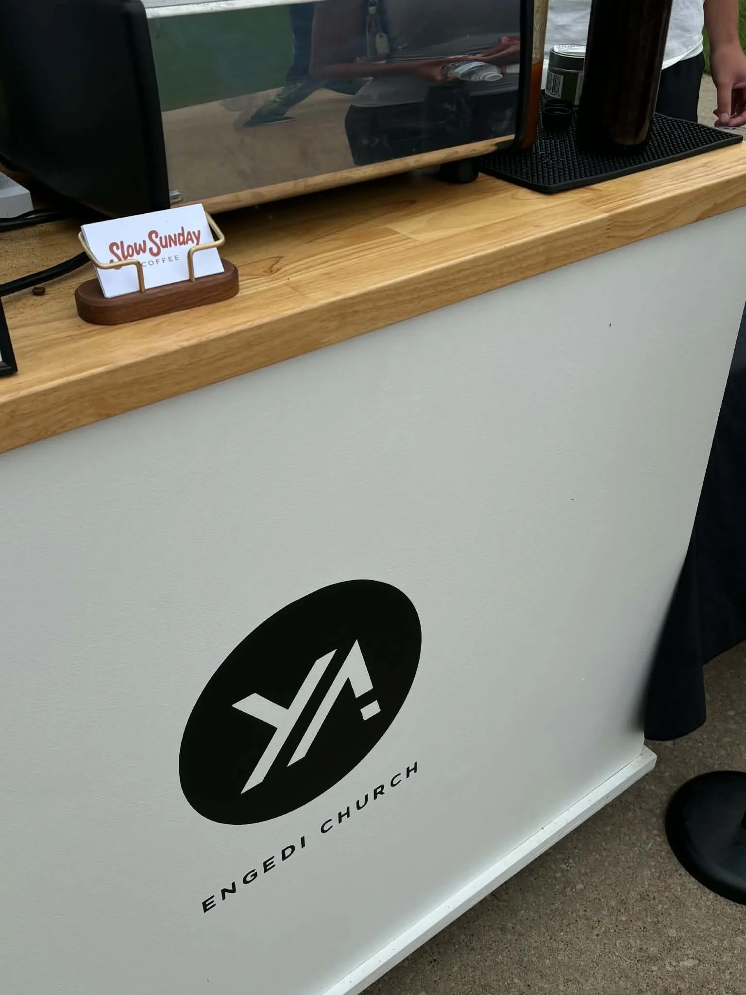 Close-up of a white food truck with black logo, a wooden countertop with a small sign reading 'Slow Sunday Coffee,' a silver cup, and part of a person's arm and leg.