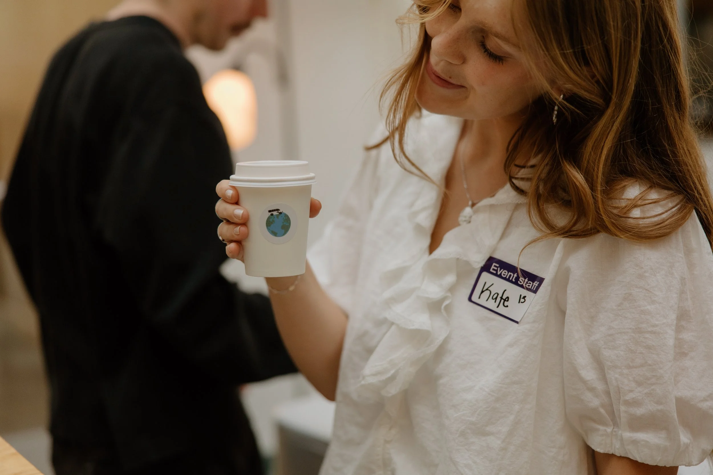 A woman with a name tag that reads 'Kate' is holding a coffee cup with a sticker of Earth on it, smiling and looking down, while a man in a dark shirt stands nearby. The background suggests an indoor event or conference.