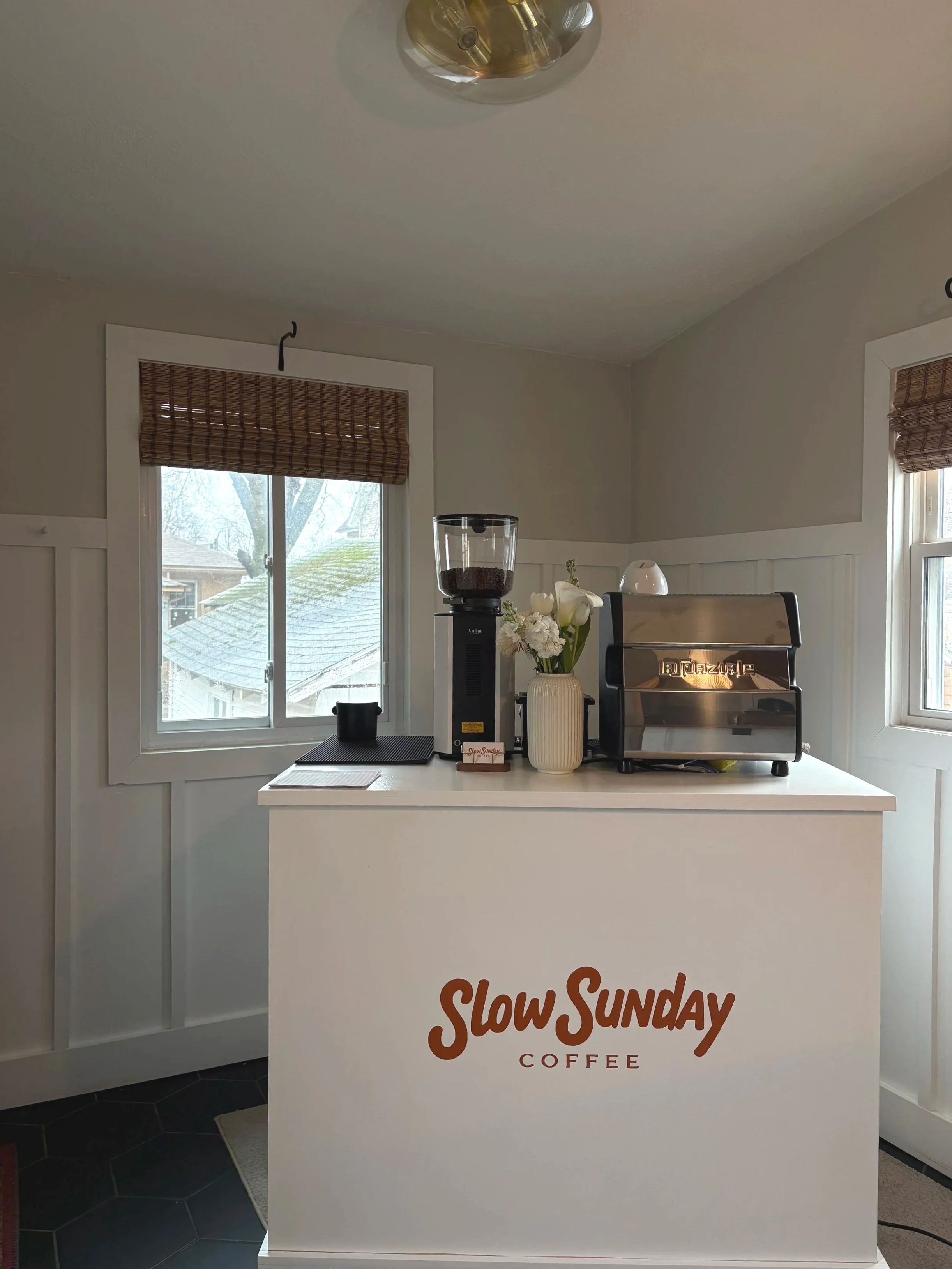 A small coffee station inside a room with white walls, featuring a white counter with 'Slow Sunday Coffee' written on it, a coffee grinder, a flower vase with white flowers, and a silver espresso machine. There are two windows with bamboo blinds.