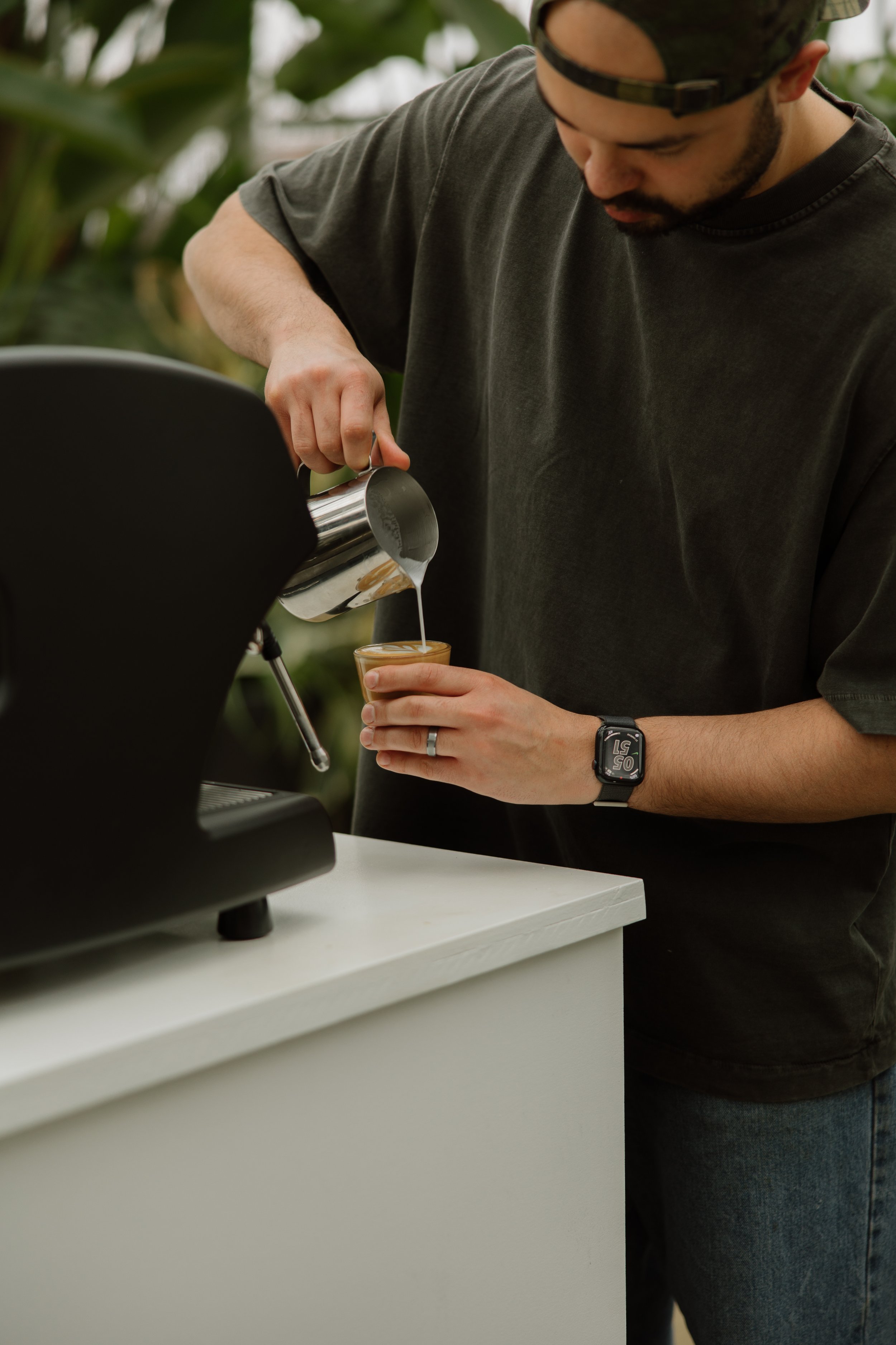 Man pouring steamed milk into a glass of coffee from a stainless steel pitcher.