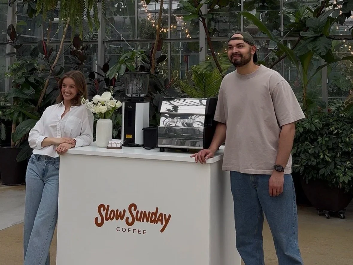 A woman and a man standing by a coffee stand inside a greenhouse with lush plants and string lights. The stand has a sign that says "Slow Sunday Coffee" and a vase with white flowers.