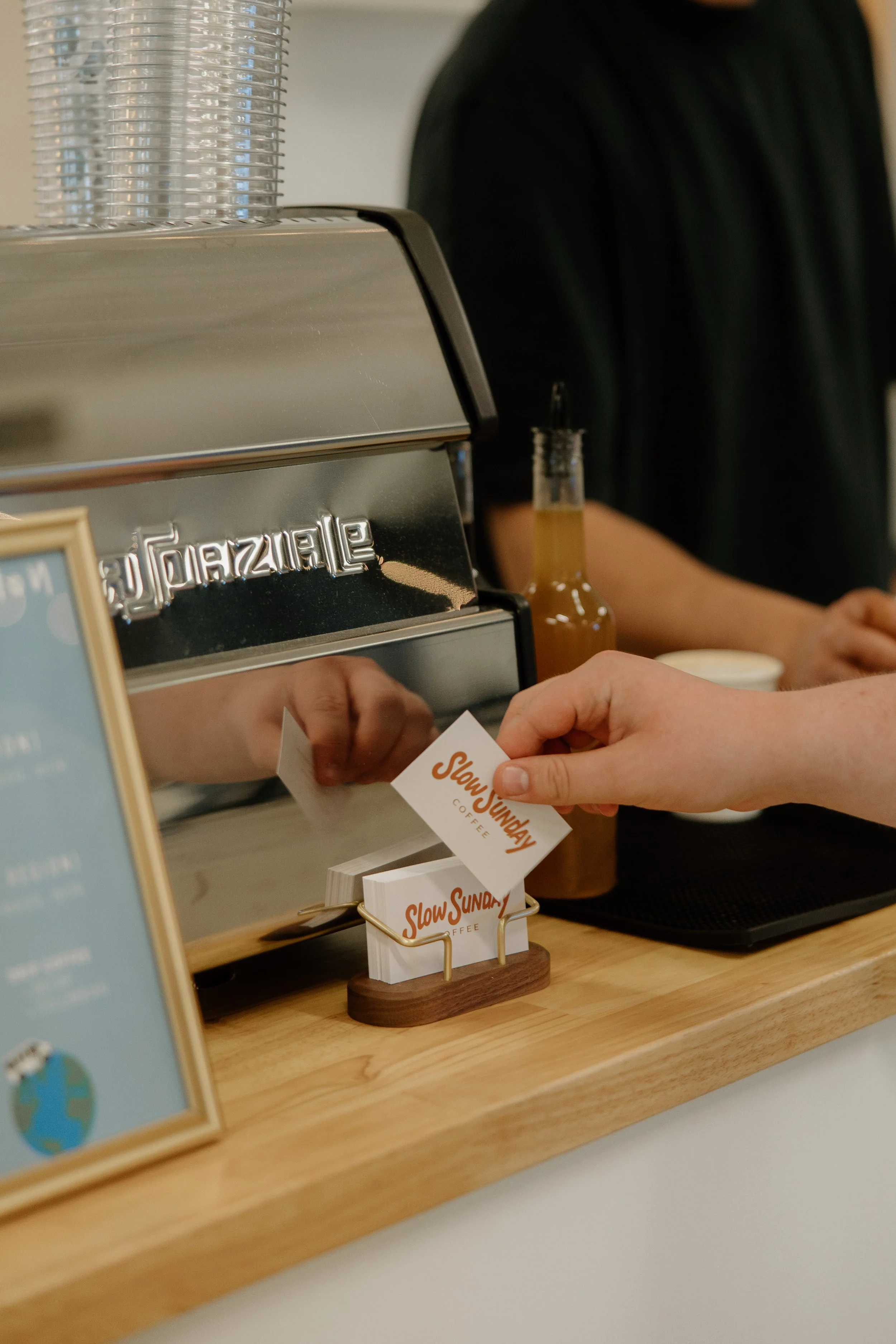 A person is seen at a coffee shop counter, with a Slow Sunday coffee sign on a small wooden display. Part of a shiny espresso machine and a clear bottle of syrup are also visible in the scene.