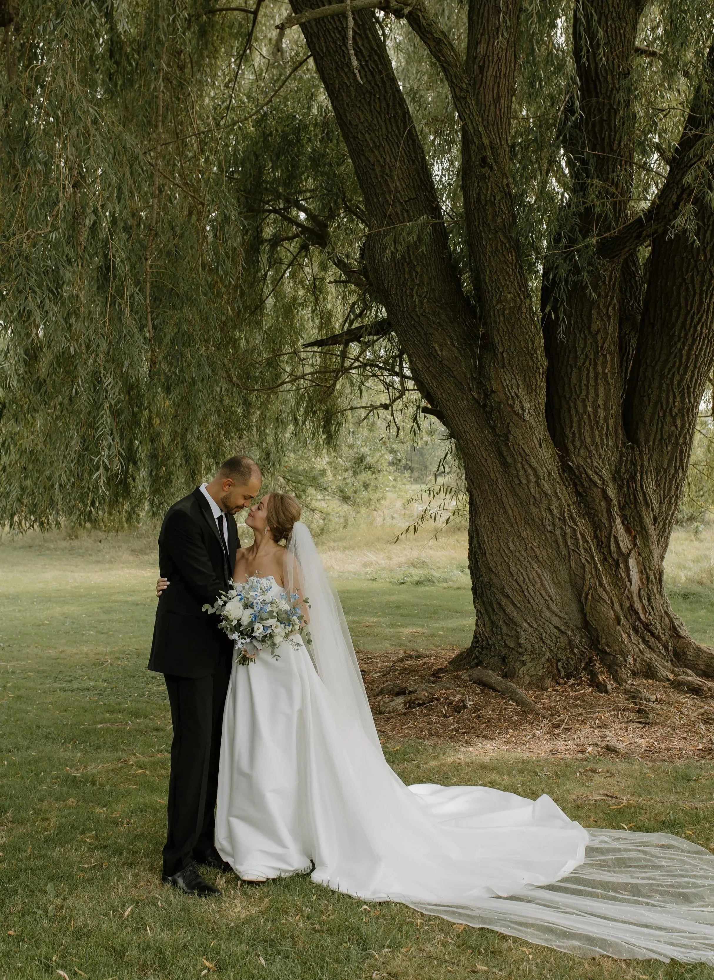 A bride and groom embrace outdoors under a large tree, with the bride holding a bouquet of white and blue flowers, both smiling and looking into each other's eyes.