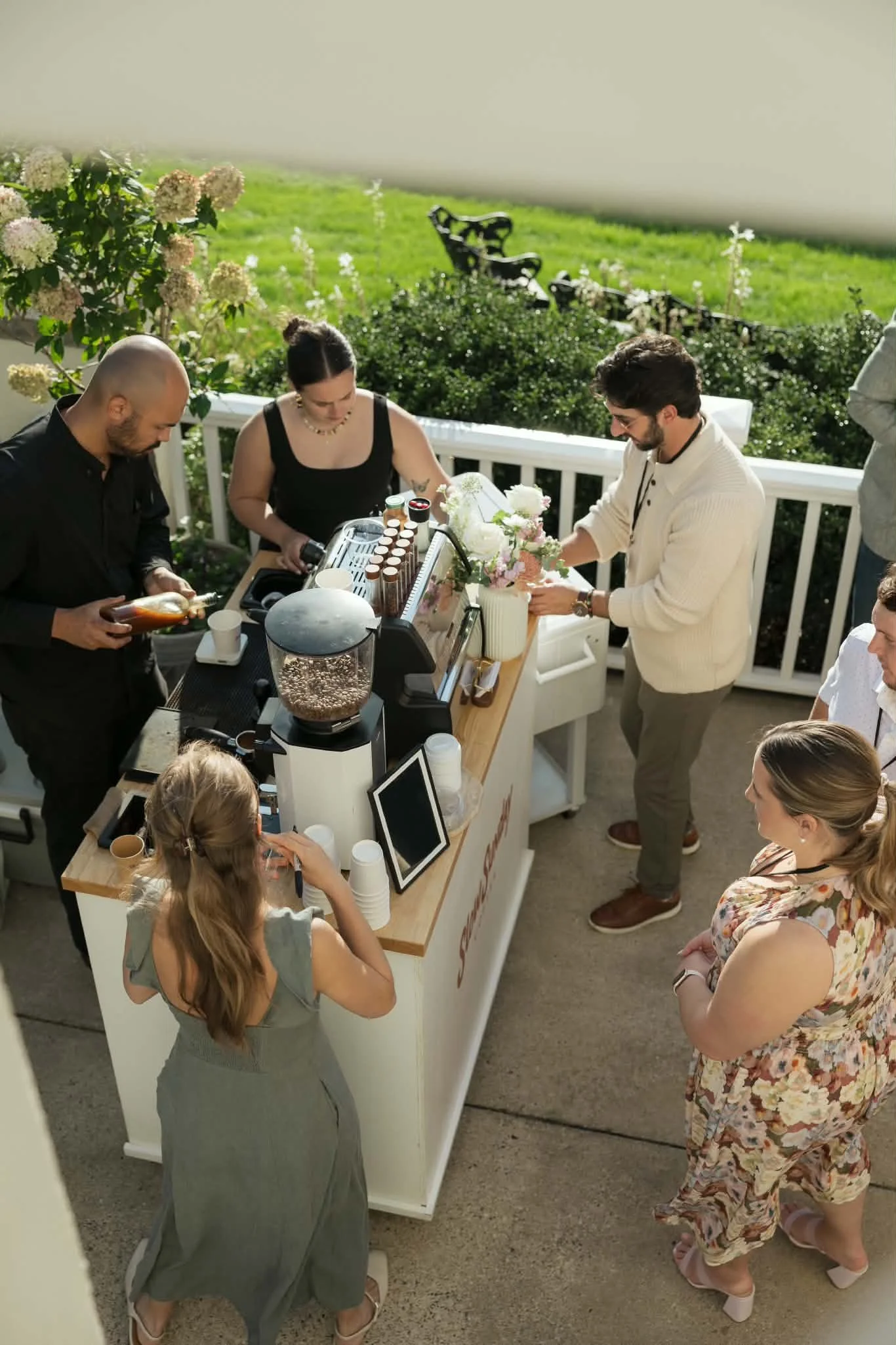 People gathered around a coffee stand outdoors, with a woman preparing drinks and others waiting in line, on a sunny day with green grass and benches in the background.