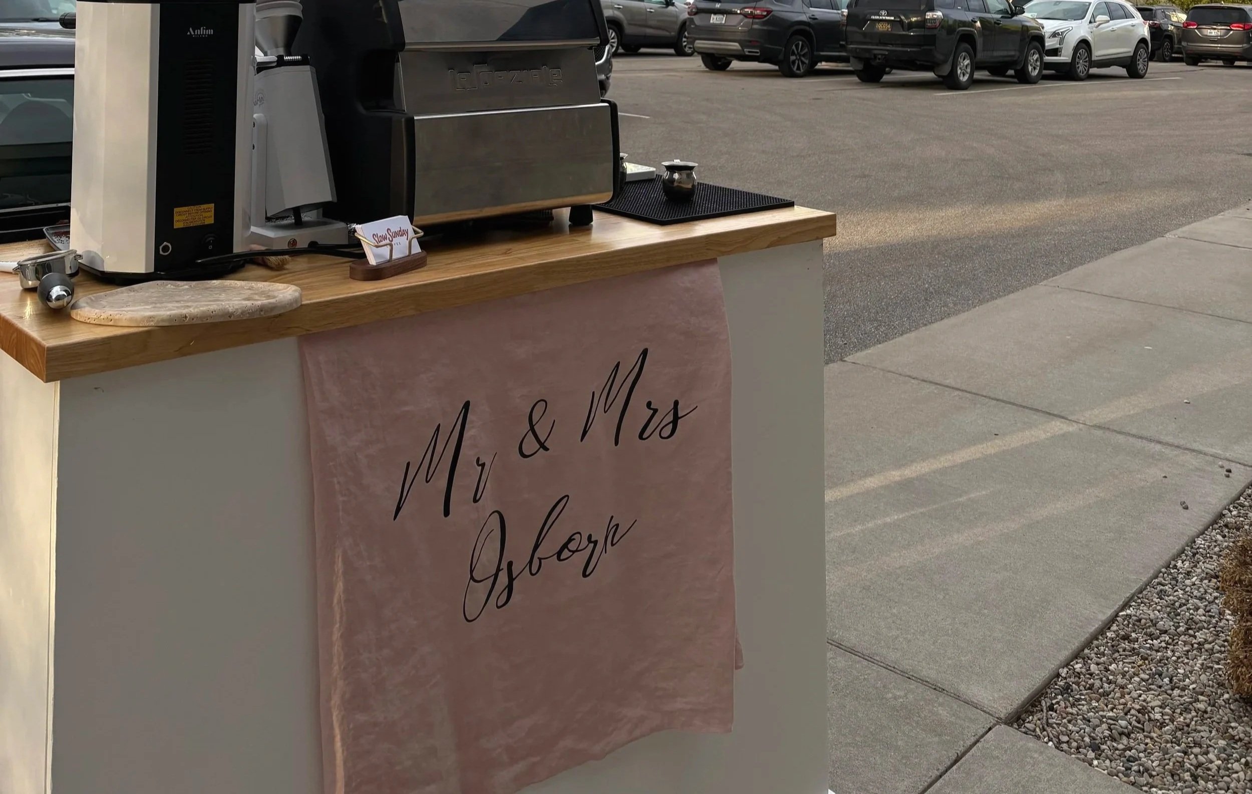 A small outdoor coffee stand with a wooden countertop, a coffee machine, and a sign that says "Mr. & Mrs. Ofor" on a pink cloth cover. Several parked cars can be seen in the background on a paved lot.