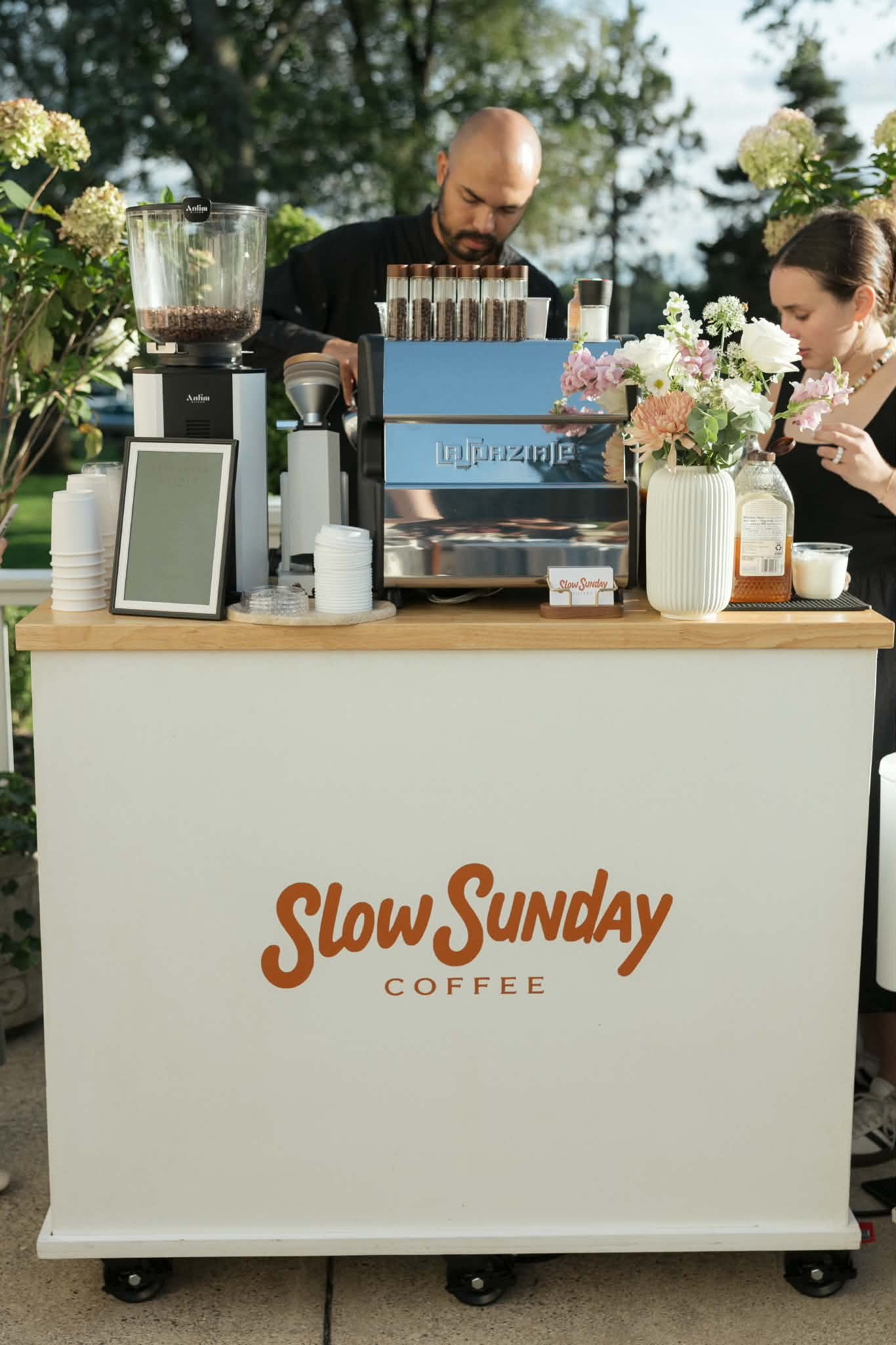 A coffee stand with a sign that says "Slow Sunday Coffee" in orange letters. The stand has flowers and coffee equipment, with a man and woman preparing drinks outdoors with trees in the background.