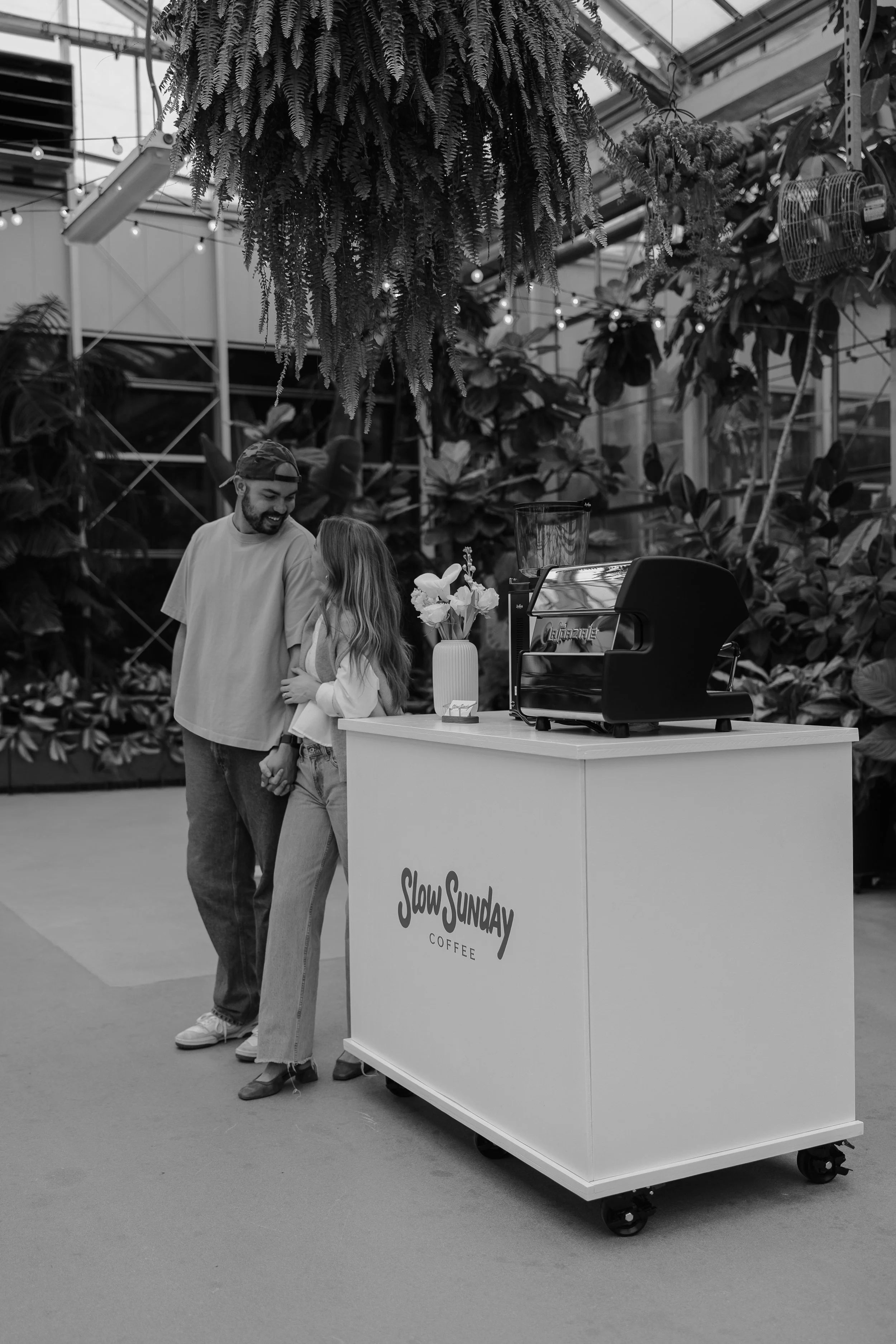 A man and a girl smiling and holding hands near a coffee cart labeled 'Slow Sunday Coffee' inside a greenhouse with hanging plants and string lights.