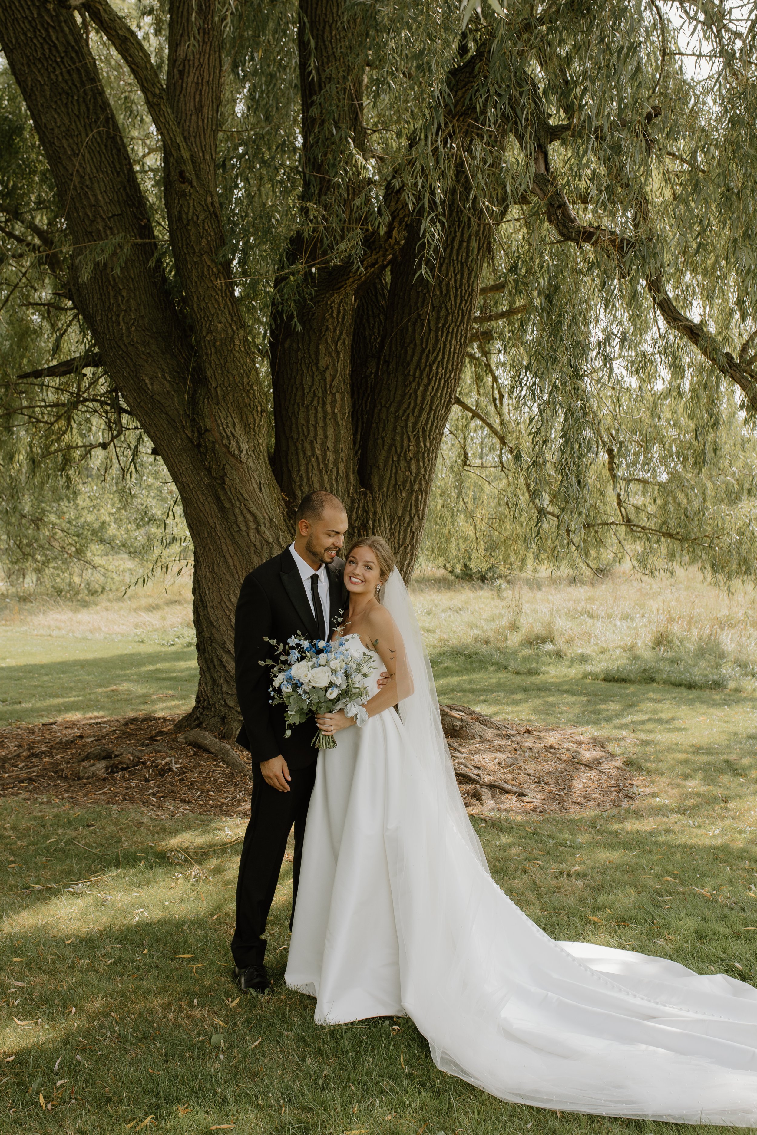 A newlywed couple standing under a large tree, smiling at each other. The bride is holding a bouquet of white and blue flowers, and wearing a white wedding dress and veil. The groom is dressed in a black suit and tie. The scene is outdoor with green grass and trees in the background.