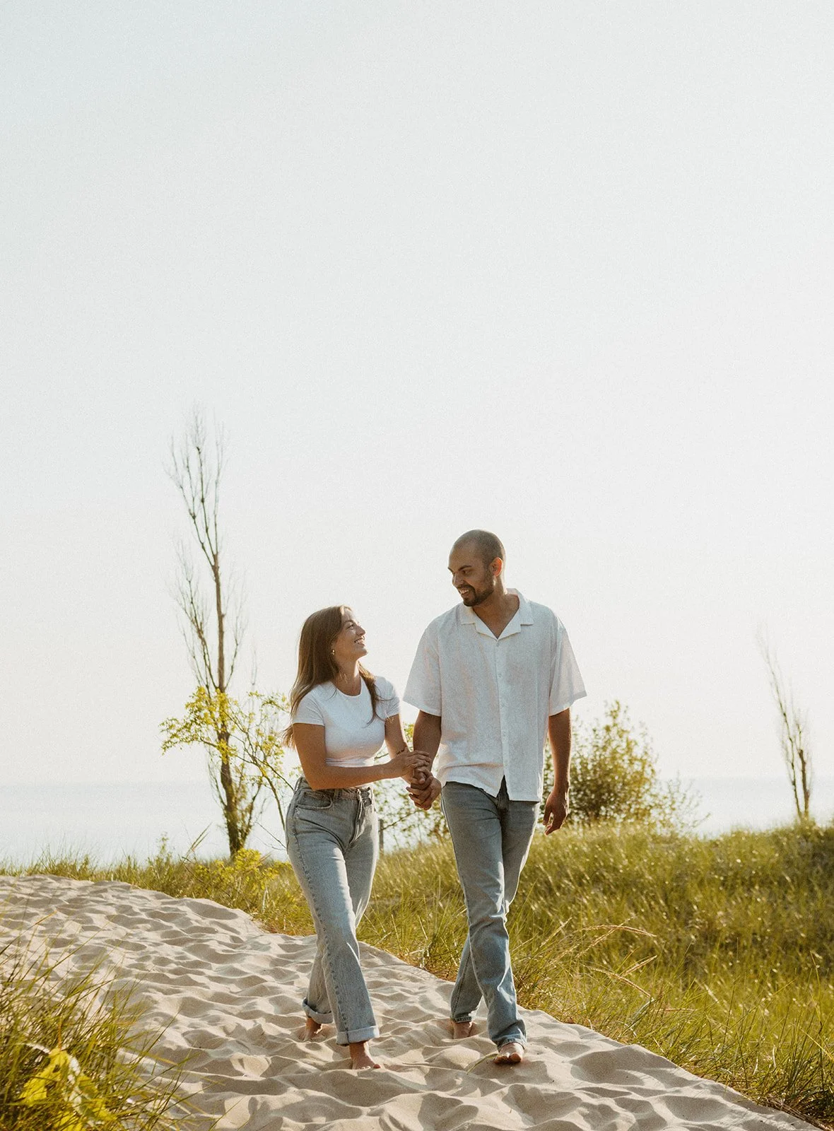 A couple walking barefoot on a sandy path through grass, holding hands and smiling at each other on a sunny day.