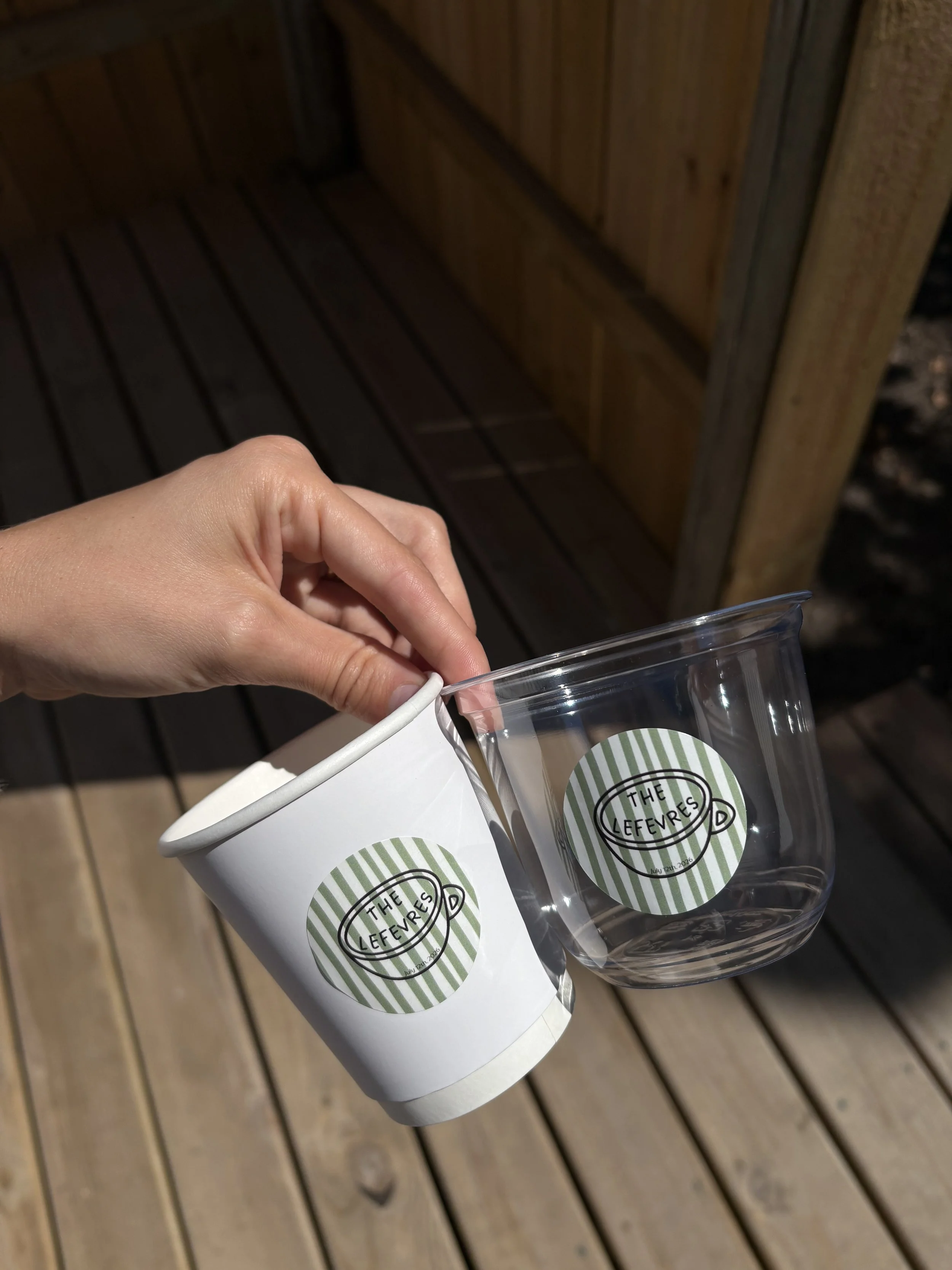 A hand holding a clear plastic cup with a paper sleeve labeled "The Lefvres" against a wooden deck background.