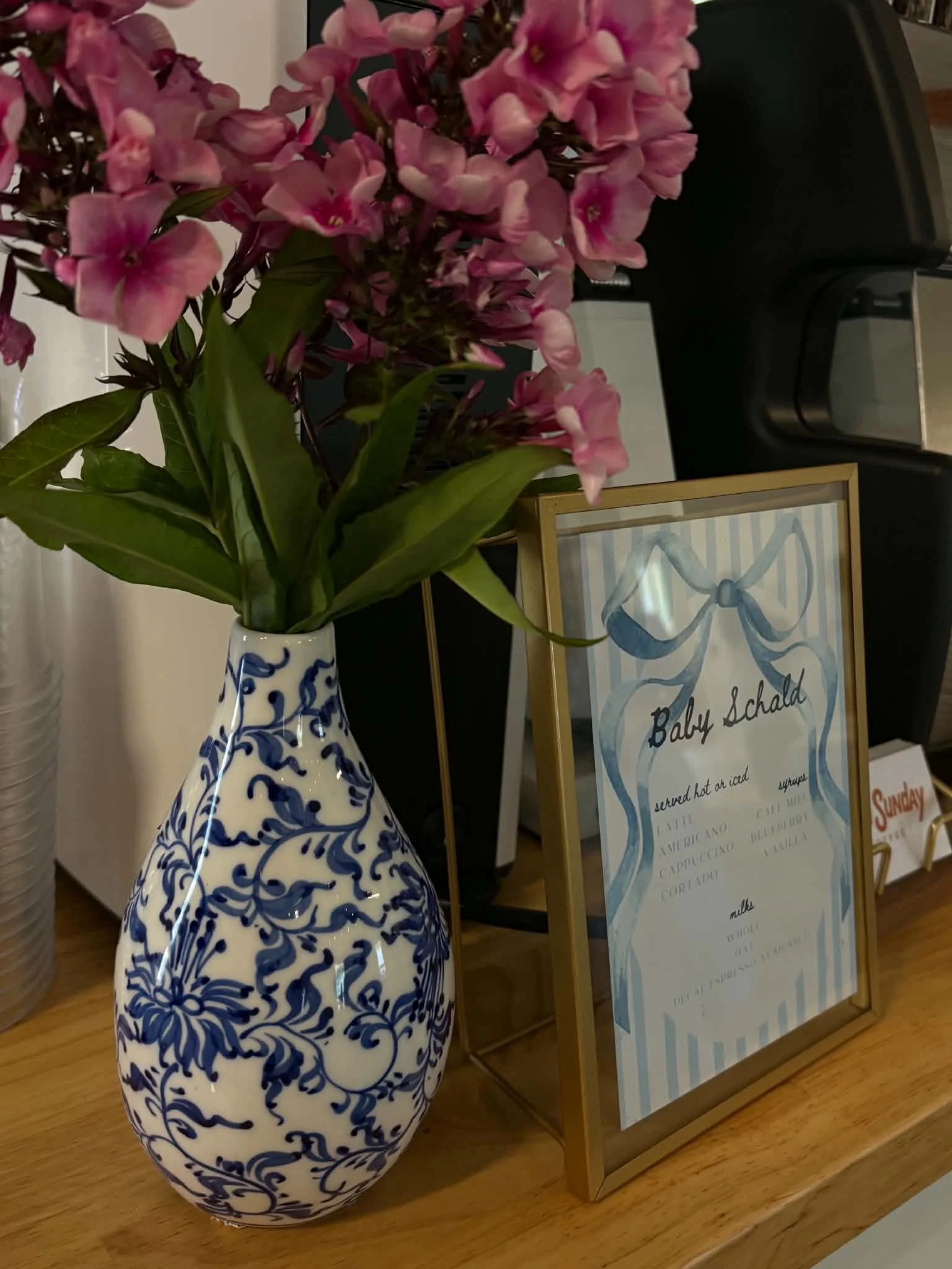 Pink flowers in a decorative blue and white vase placed on a wooden surface, with a framed sign that reads 'Baby Schald served hot or iced' and a black coffee machine in the background.