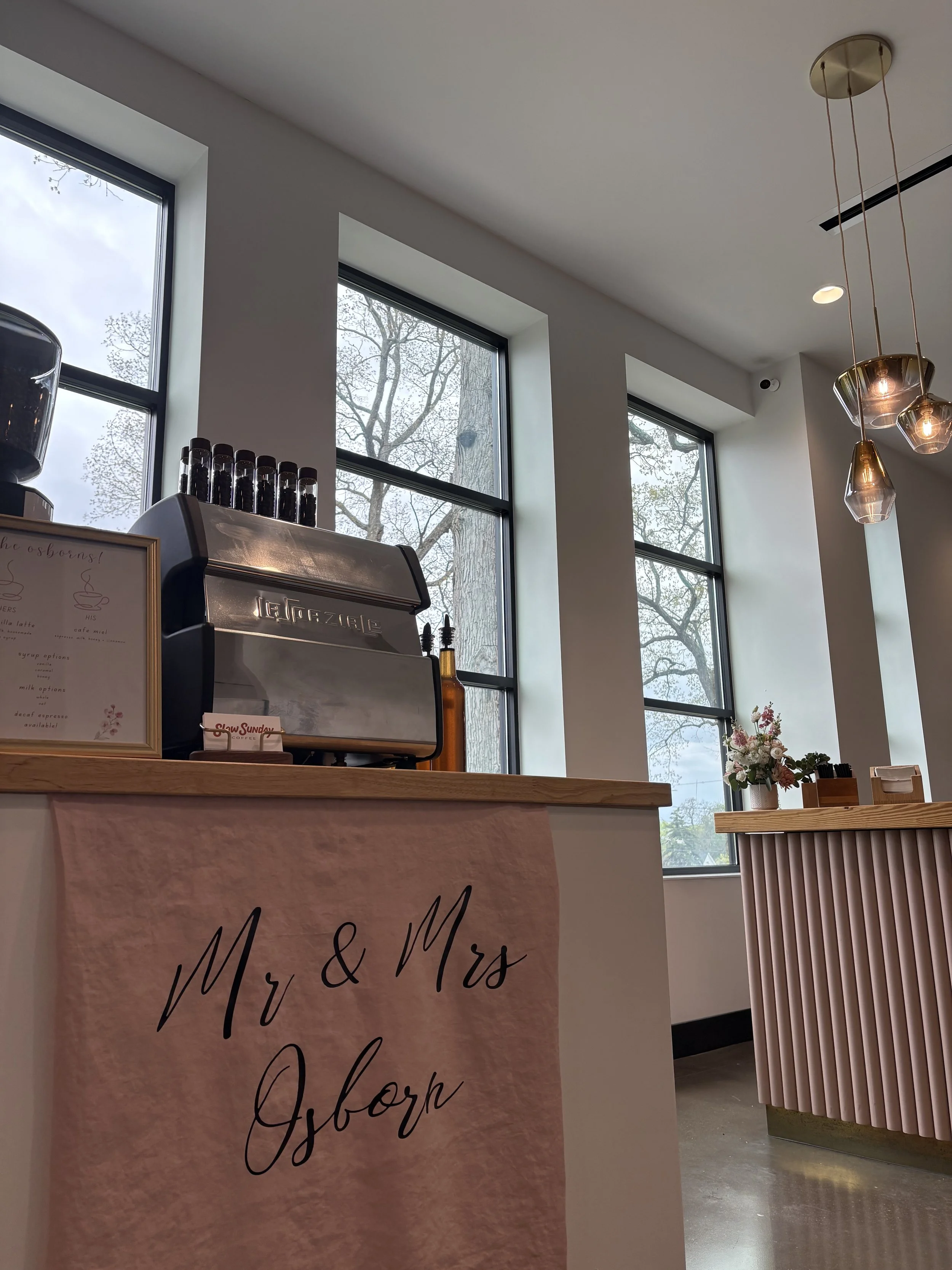 Interior of a cafe with large windows, a coffee machine on a counter, a pink banner with 'Mr. & Mrs.' written on it, and a wooden table with flowers. Elegant hanging light fixtures are visible.