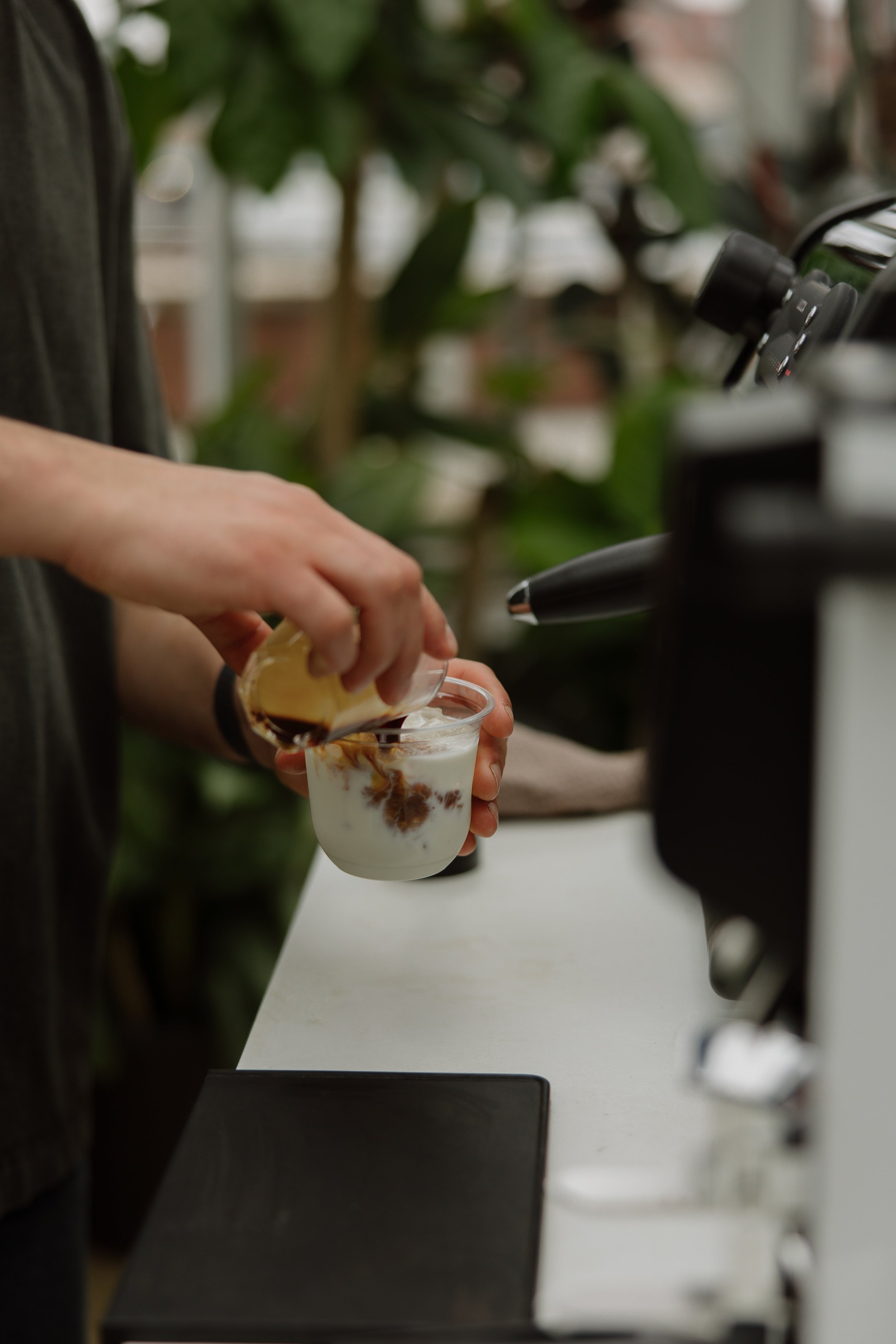 A person making an iced coffee or similar beverage with ice, milk, and coffee at a coffee shop or cafe, with greenery in the background.