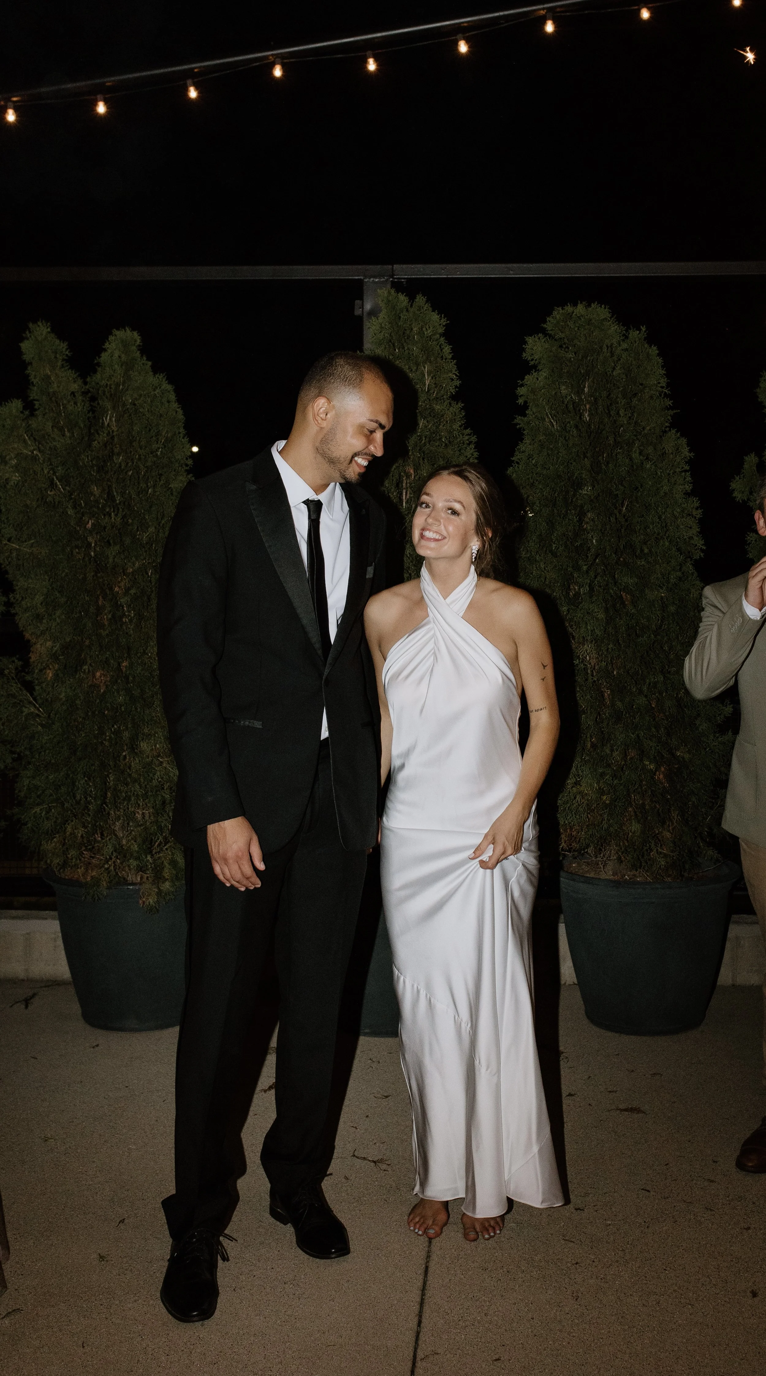 A man in a black tuxedo and a woman in a white satin dress are smiling and looking at each other at night, surrounded by potted evergreen plants, with string lights overhead.