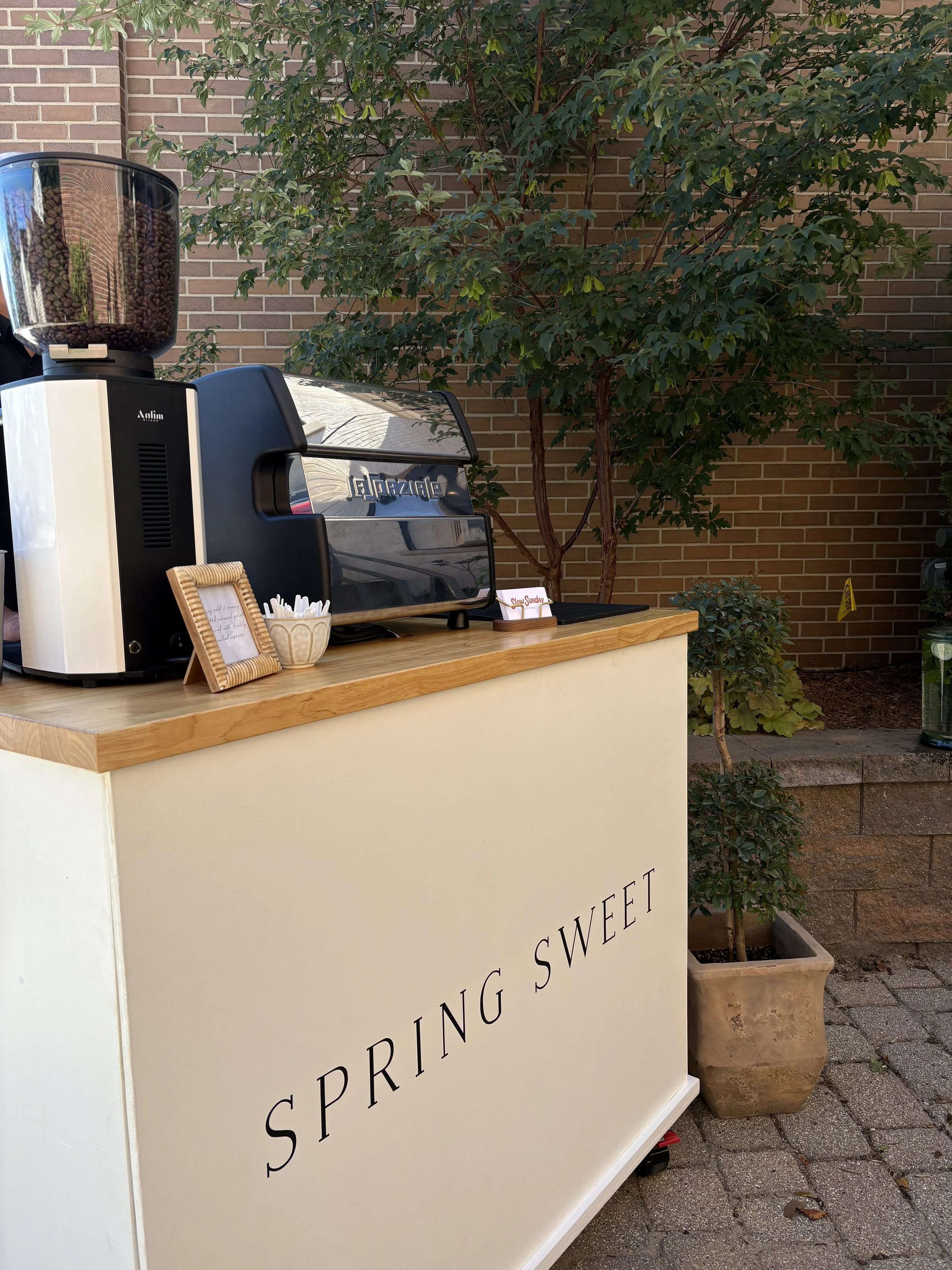 A coffee and espresso stand with a white counter, a coffee grinder, a commercial espresso machine, and a small framed sign, outdoors near a brick wall and green trees.