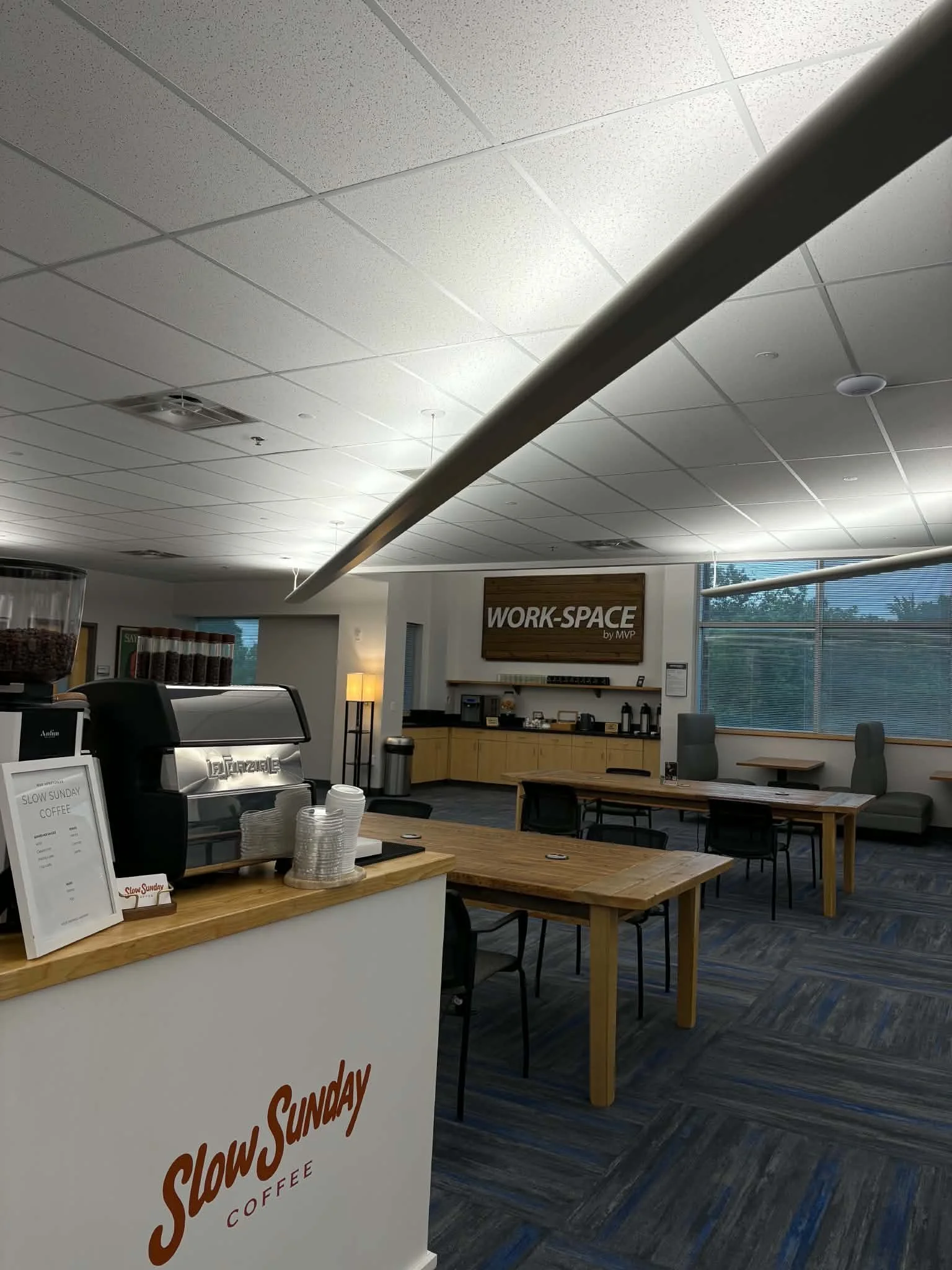 Interior of a coffee shop with wooden tables and chairs, a coffee counter, and a sign that reads "Slow Sunday Coffee". There are large windows with blinds and modern lighting fixtures.