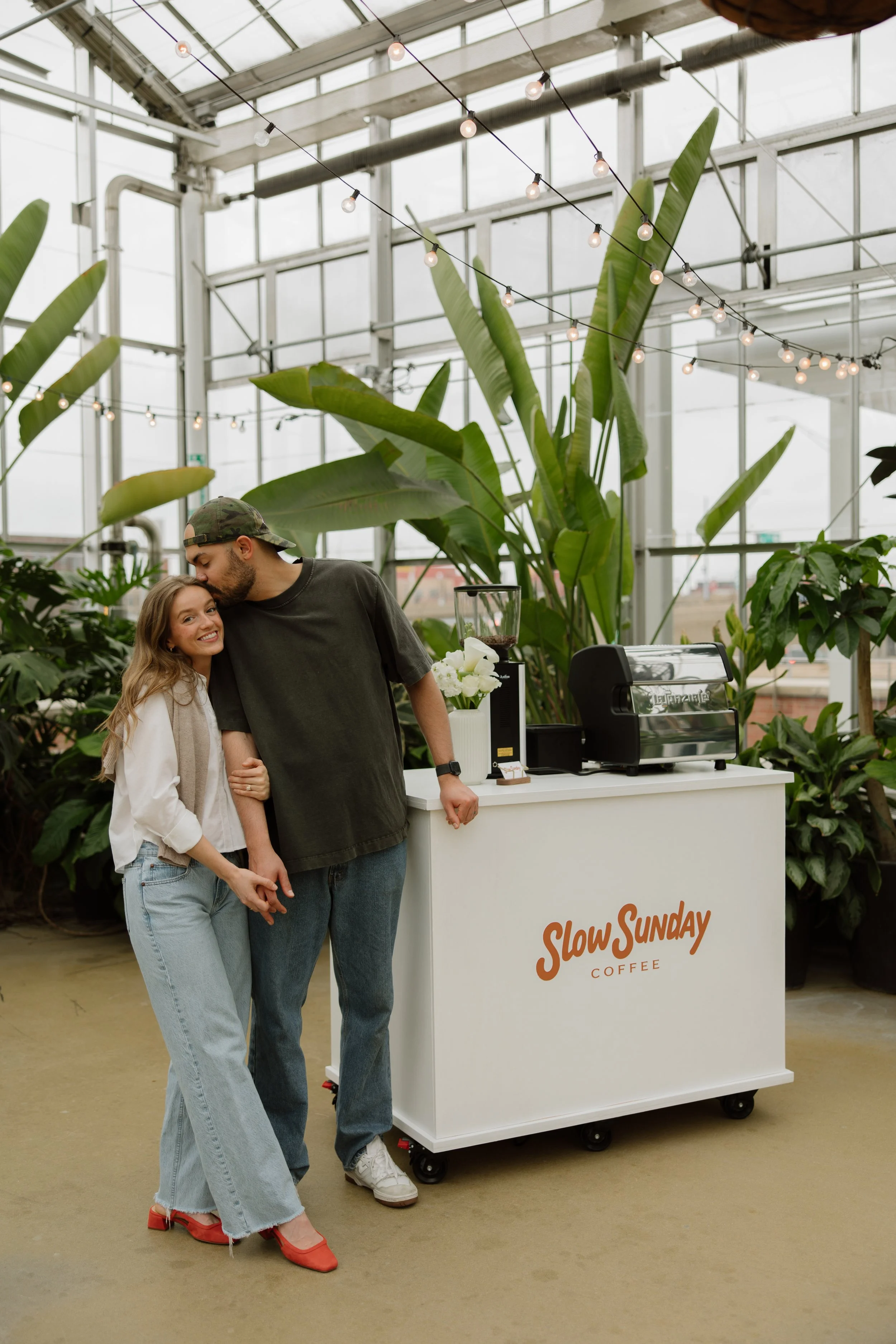 A smiling woman and a man hugging inside a greenhouse or plant shop with large tropical plants, string lights overhead, and a coffee stand labeled 'Slow SUNDAY COFFEE' in the background.