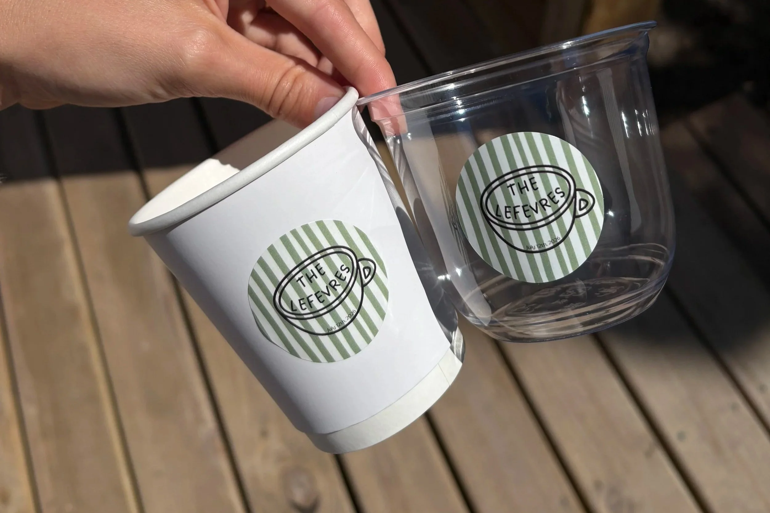A hand holding a clear plastic container with a sticker that reads 'The Leferves' and a striped pattern, placed on a wooden surface.