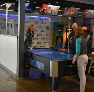 Three women playing air hockey at an arcade, with multiple arcade game screens visible in the background.