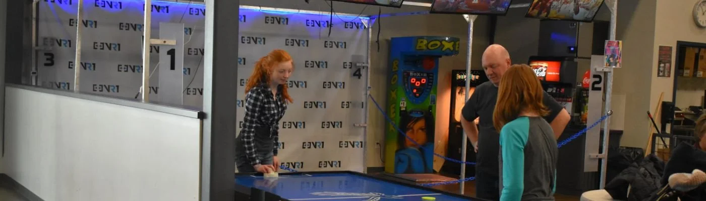 Three people are playing air hockey at an arcade. Two women and one man are around the air hockey table, which has a blue surface, while a girl with red hair in a checkered shirt is on the other side of the table. The background shows an arcade game and a Coca-Cola vending machine.