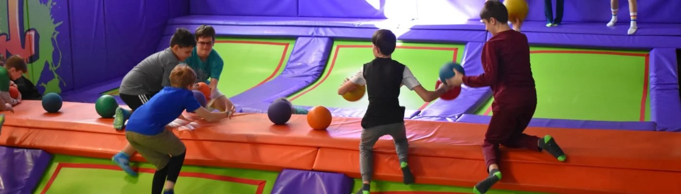 Children playing with sports balls on a padded, colorful indoor trampoline area.