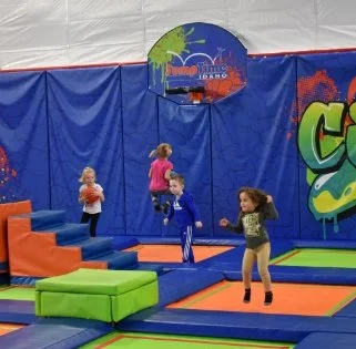 Children playing on indoor trampoline and padded play area with blue walls and colorful equipment.