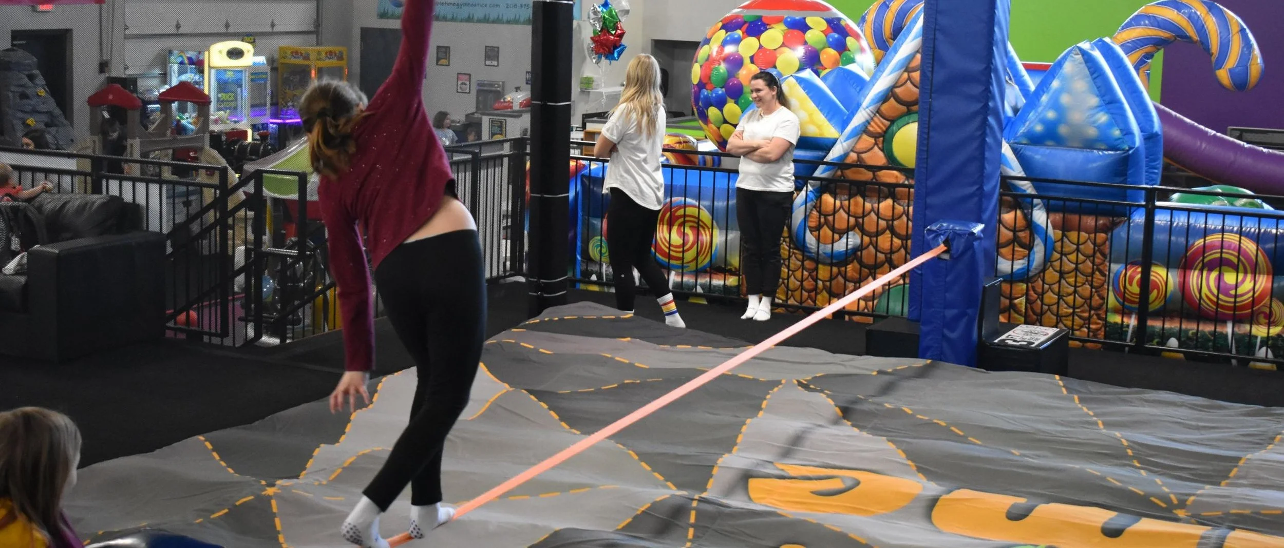 An indoor amusement area with a girl in a maroon sweater jumping on a slackline, two women in white shirts observing, and colorful inflatable play structures in the background.