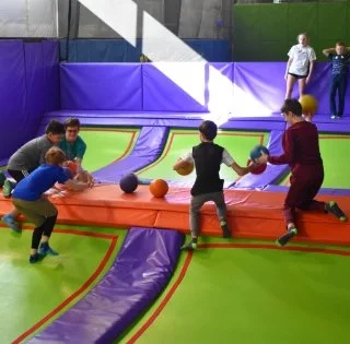Children and adults playing dodgeball inside an indoor trampoline park with padded walls and colorful mats.