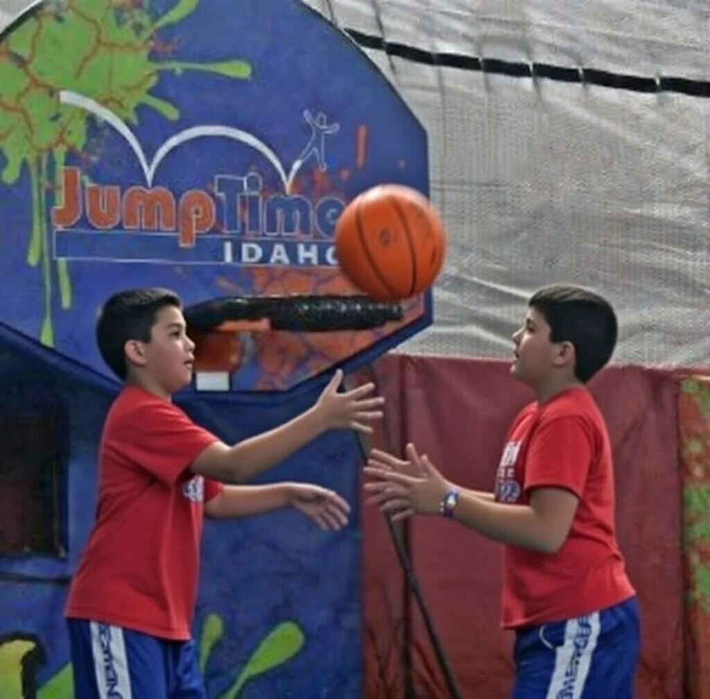 Two boys in red shirts playing basketball in an indoor bounce house with a Jump Time Idaho sign in the background.
