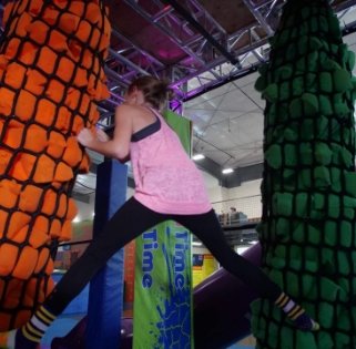 A girl climbing an obstacle course with foam cylinders shaped like rocks, one orange and one green, in an indoor play area.