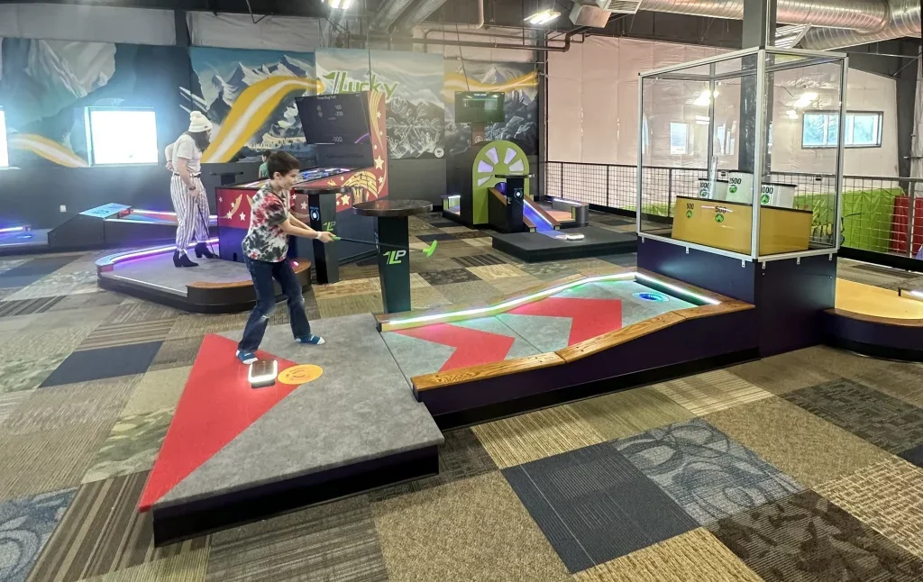 Indoor mini-golf course with colorful, themed holes. Two children are playing, and an adult is watching in the background. The course features various obstacles, ramps, and bright lighting.