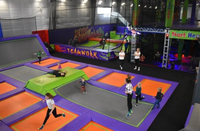 Indoor trampoline park with multiple interconnected trampolines, padded areas in orange and green, children bouncing, and adults supervising, with colorful walls and a