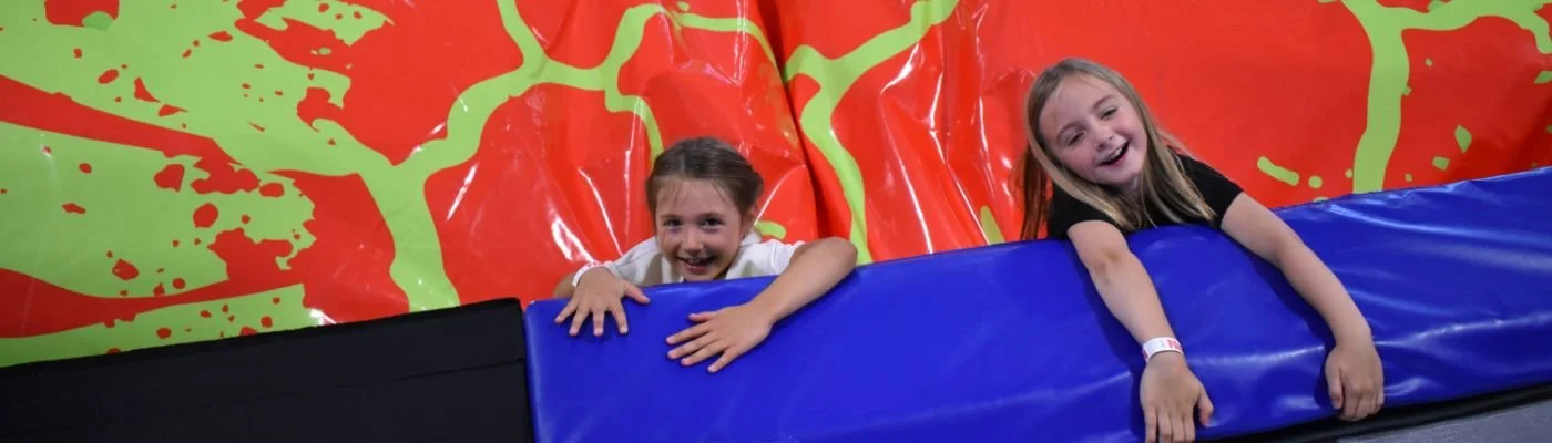 Two young girls smiling and playing on an indoor trampoline with a colorful red, green, and yellow padded wall in the background.