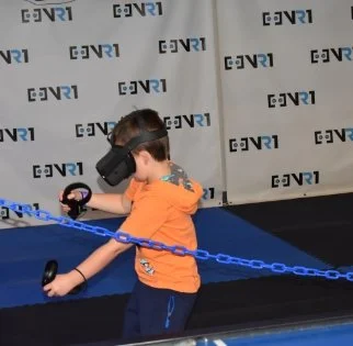 A young boy wearing an orange shirt and virtual reality goggles, holding VR controllers, standing inside a VR attraction with a blue chain barrier.