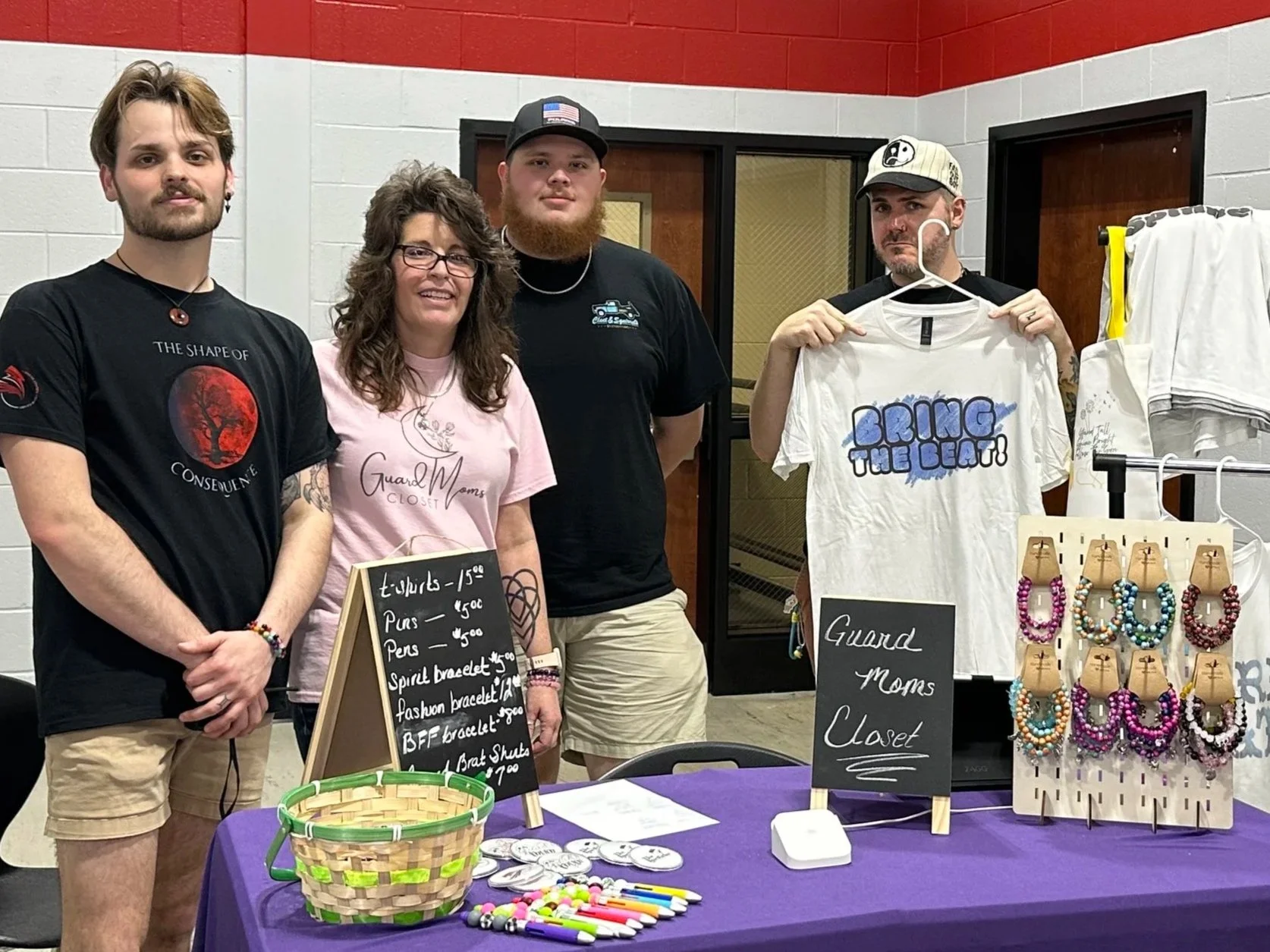 A group of four people standing behind a table at a craft fair, displaying jewelry and t-shirts. The table has a purple cloth and various jewelry items on display, including beaded bracelets. There are signs with prices and a small basket, with a backdrop of a wall with red and gray bricks.