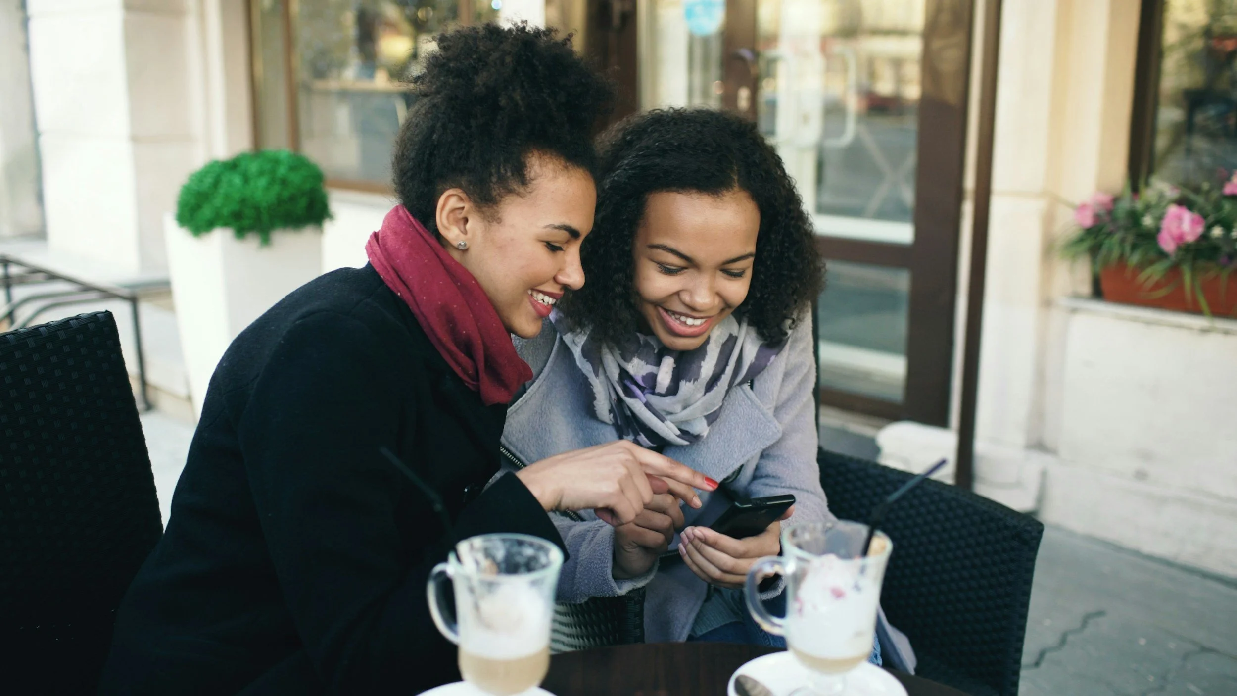 Two women sharing drinks and looking at a smartphone outside a cafe, smiling