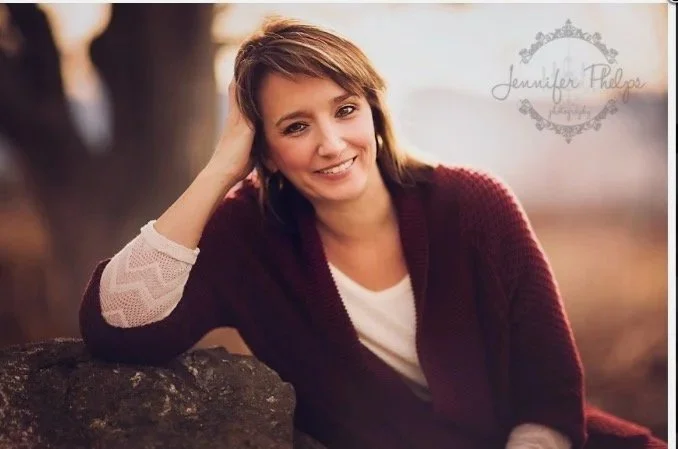 A woman with brown hair smiling, leaning on a rock outdoors during sunset. She is wearing a white top with a maroon cardigan.