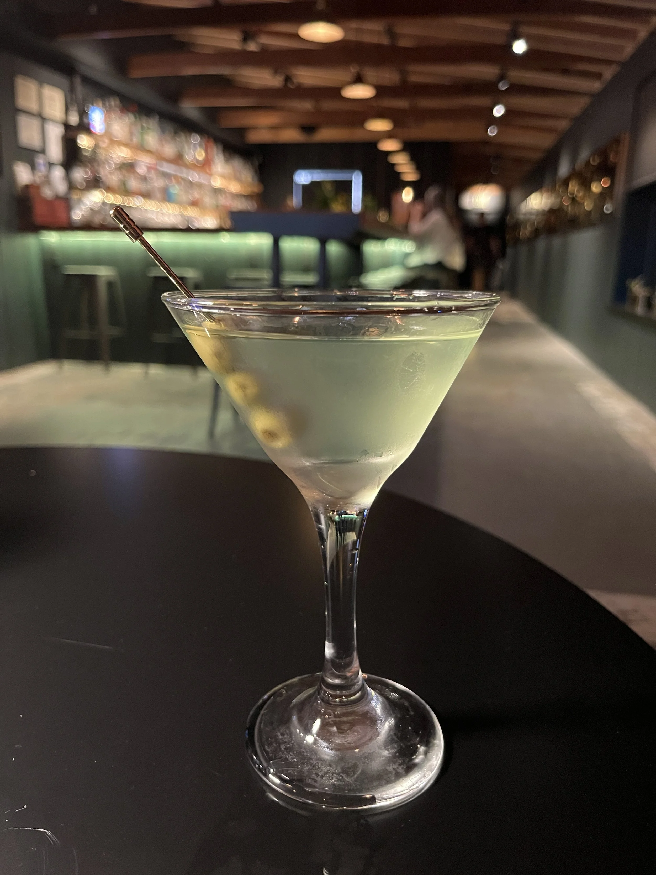 A green cocktail in a stemmed martini glass with a metal straw, set on a black table in a dimly lit bar with wooden ceiling beams and blurred background.