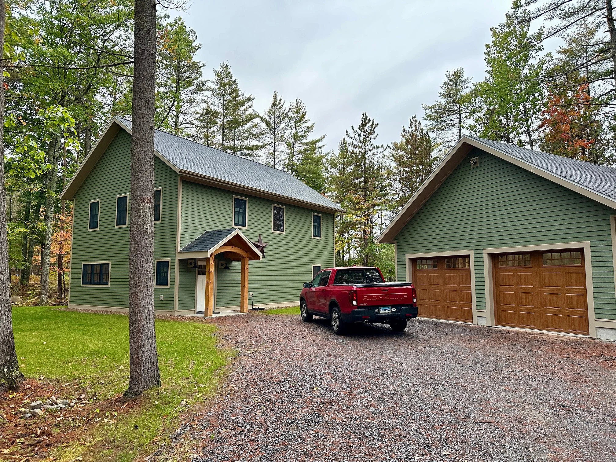 Green house with brown garage doors and a red truck parked in the driveway, surrounded by tall trees with some autumn foliage, and cloudy sky.