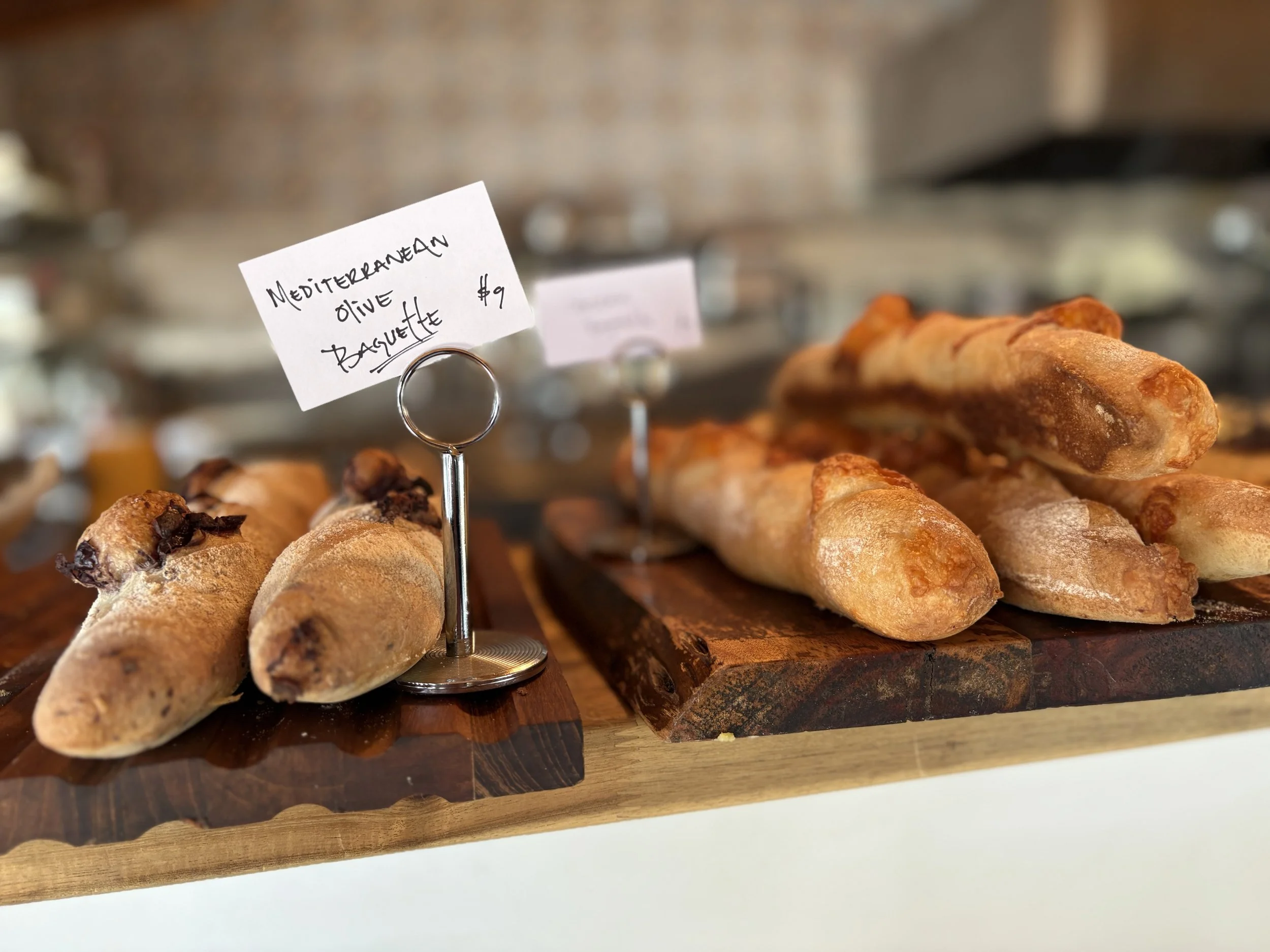 Close-up of breadsticks with a handwritten sign that reads 'Mediterranean Olive Basket' on a stand, placed on a wooden serving board.
