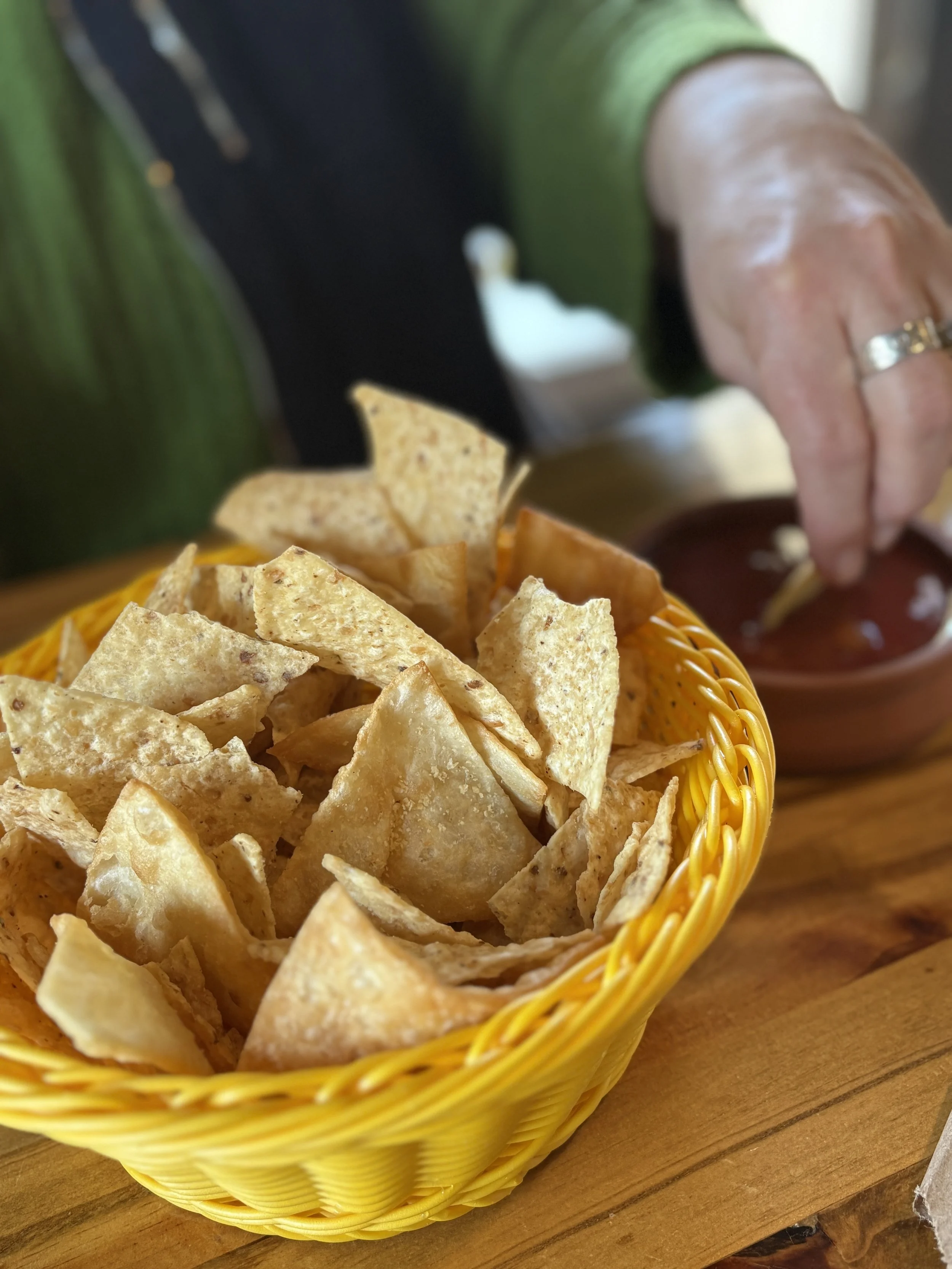 A yellow basket filled with tortilla chips on a wooden table, with a person reaching for some and a bowl of salsa in the background.