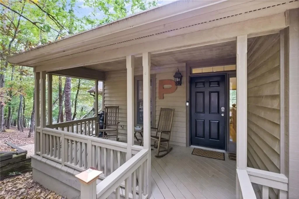 View of a house porch with two wooden rocking chairs, a black front door, a lantern-style porch light, and a decor piece with the letter 'P' on the wall, surrounded by trees in a wooded area.