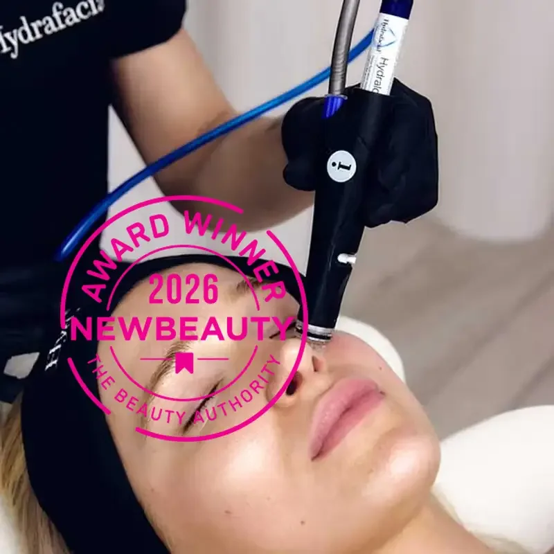 A woman receiving a cosmetic or dermatological treatment with a device held by a professional wearing black gloves. A pink circular badge overlay reads "Award Winner 2026 NewBeauty The Beauty Authority."