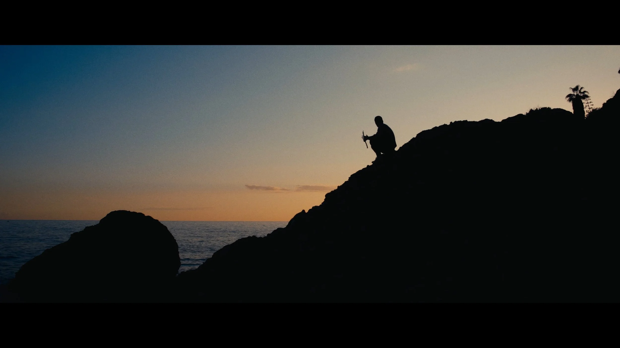 Silhouette of a person playing the flute on a rocky shoreline during sunset.