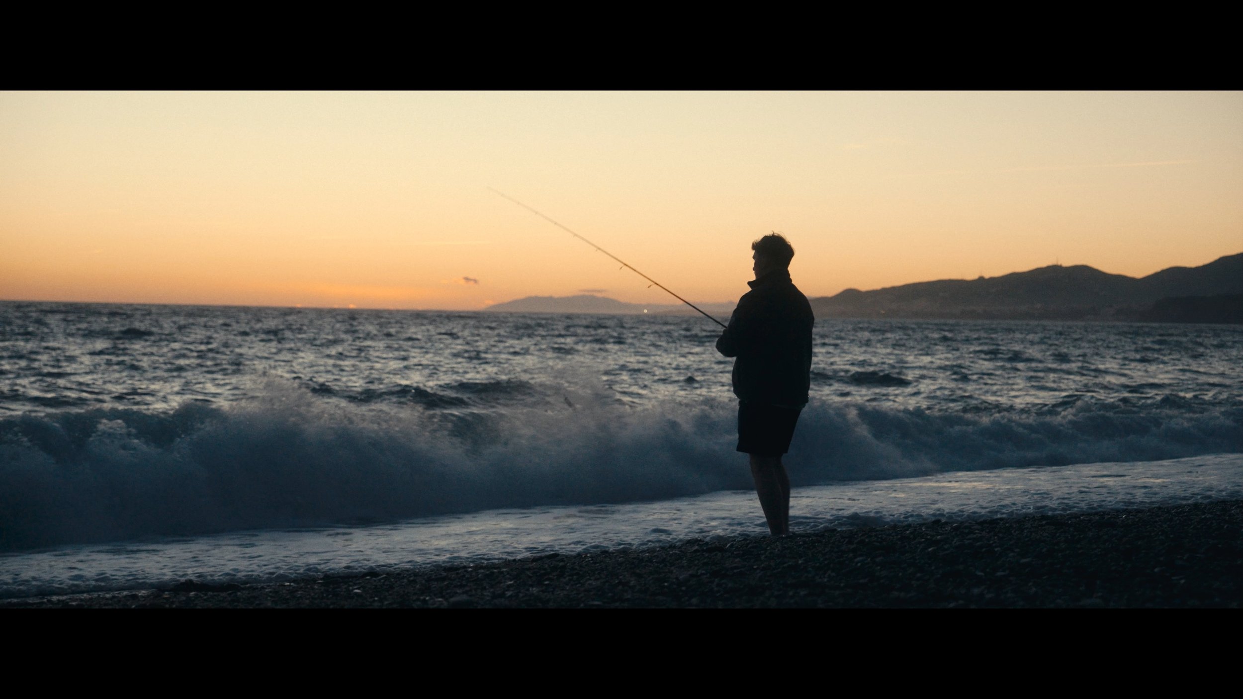 A person standing on a beach at sunset, fishing with a rod.