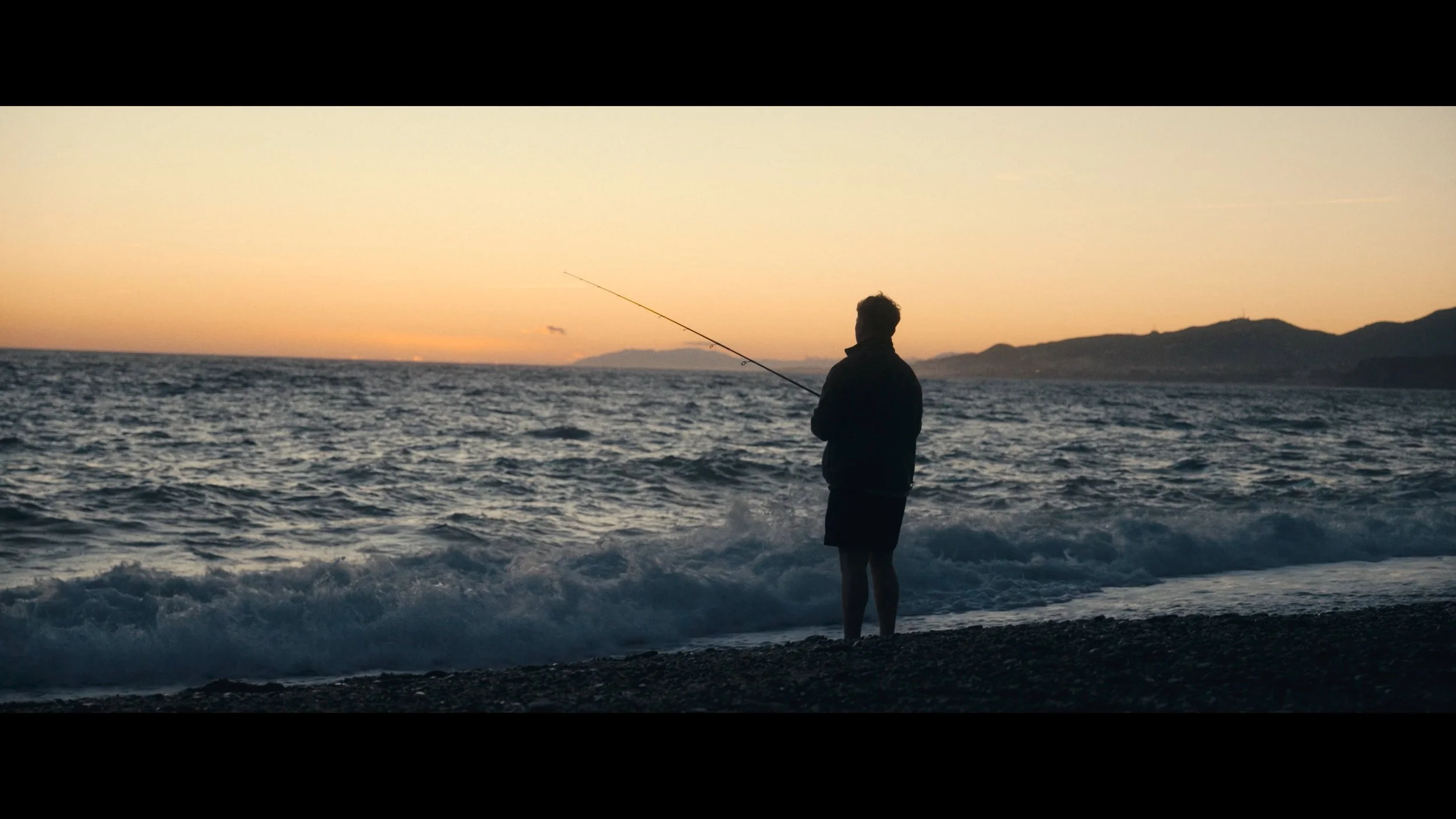A person standing on a rocky beach at sunset, fishing in the ocean with mountains in the distance.